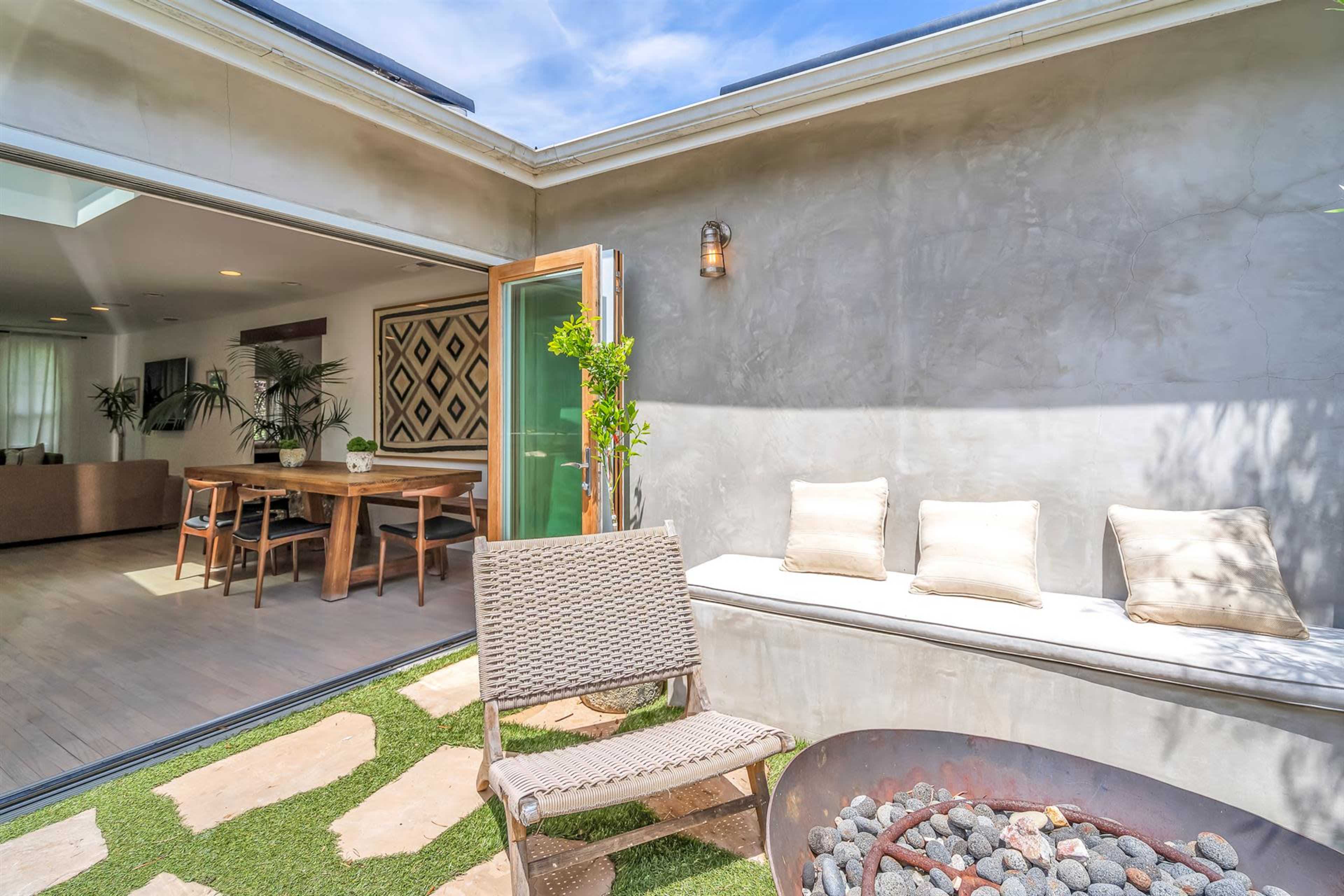 The image shows a modern outdoor seating area with a fire pit, surrounded by grass and accessible from an indoor dining space.