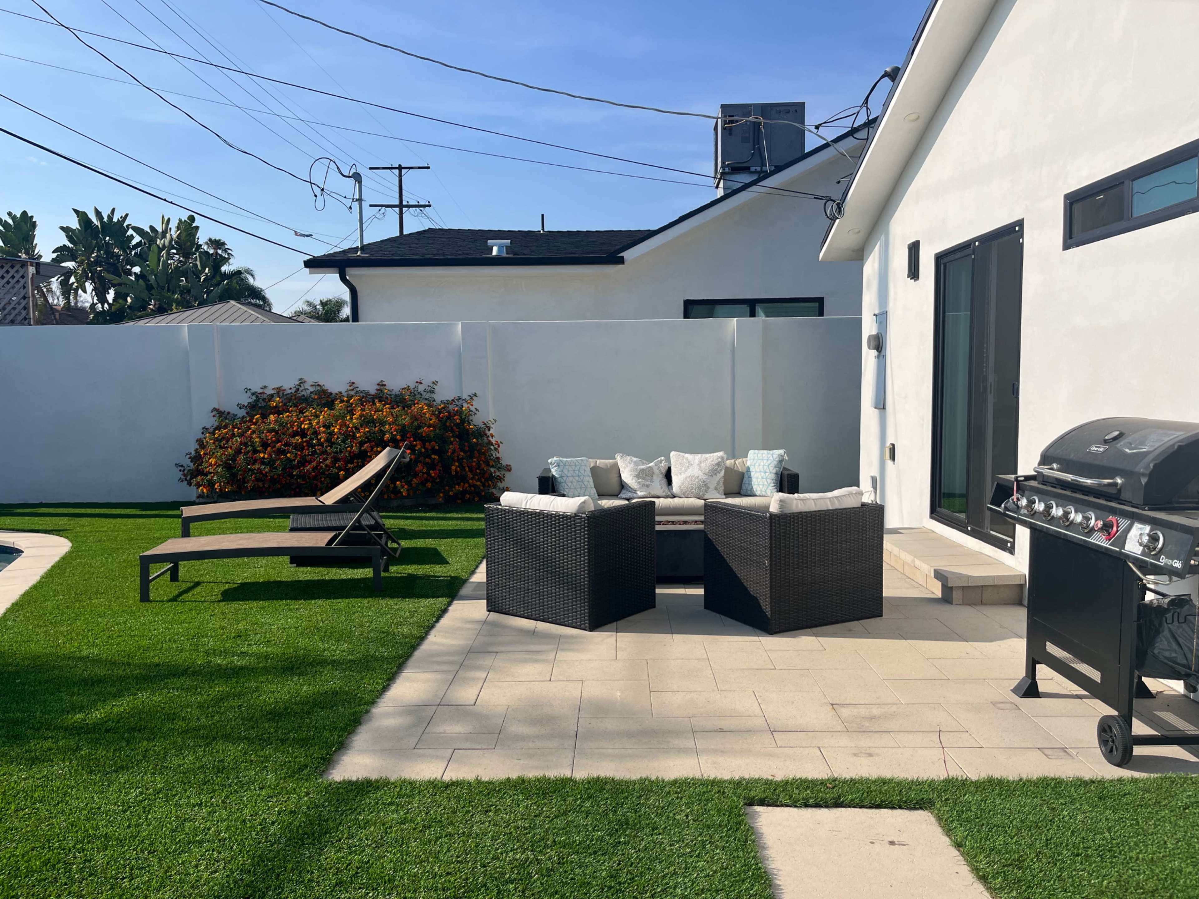 A patio area features a seating arrangement with cushions, a lounge chair, and a barbecue grill surrounded by greenery and a white wall.