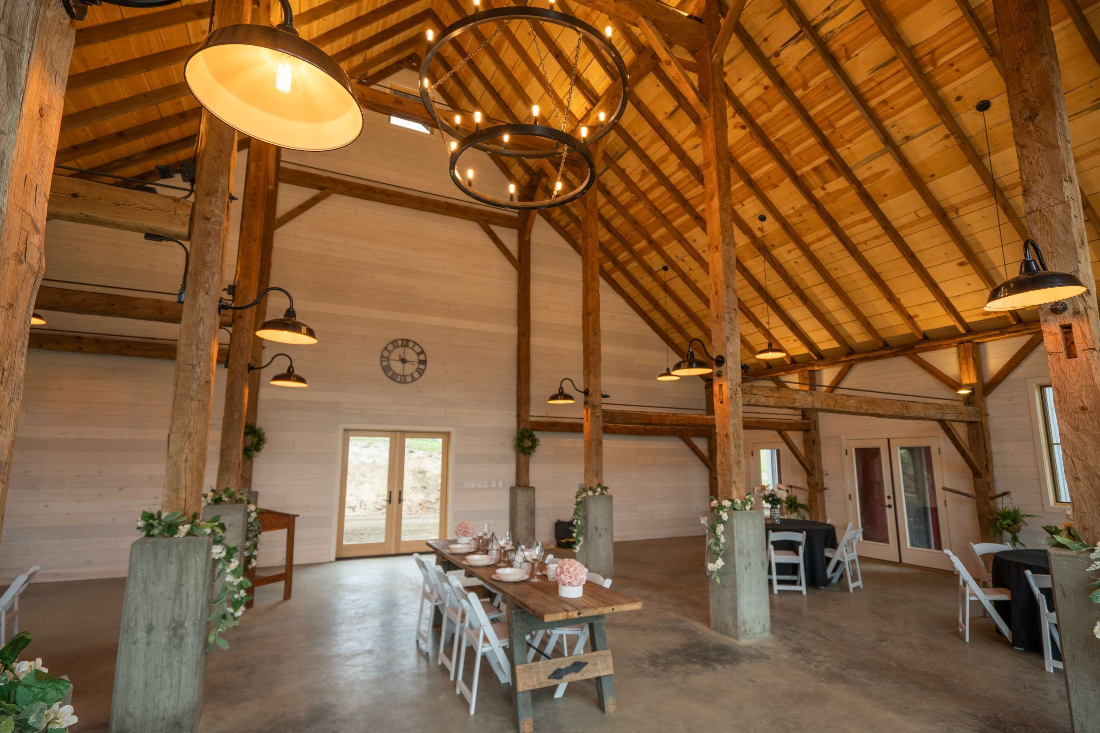 The interior of a spacious, wooden barn with a high ceiling, a long dining table set for an event, and decorative lighting fixtures.