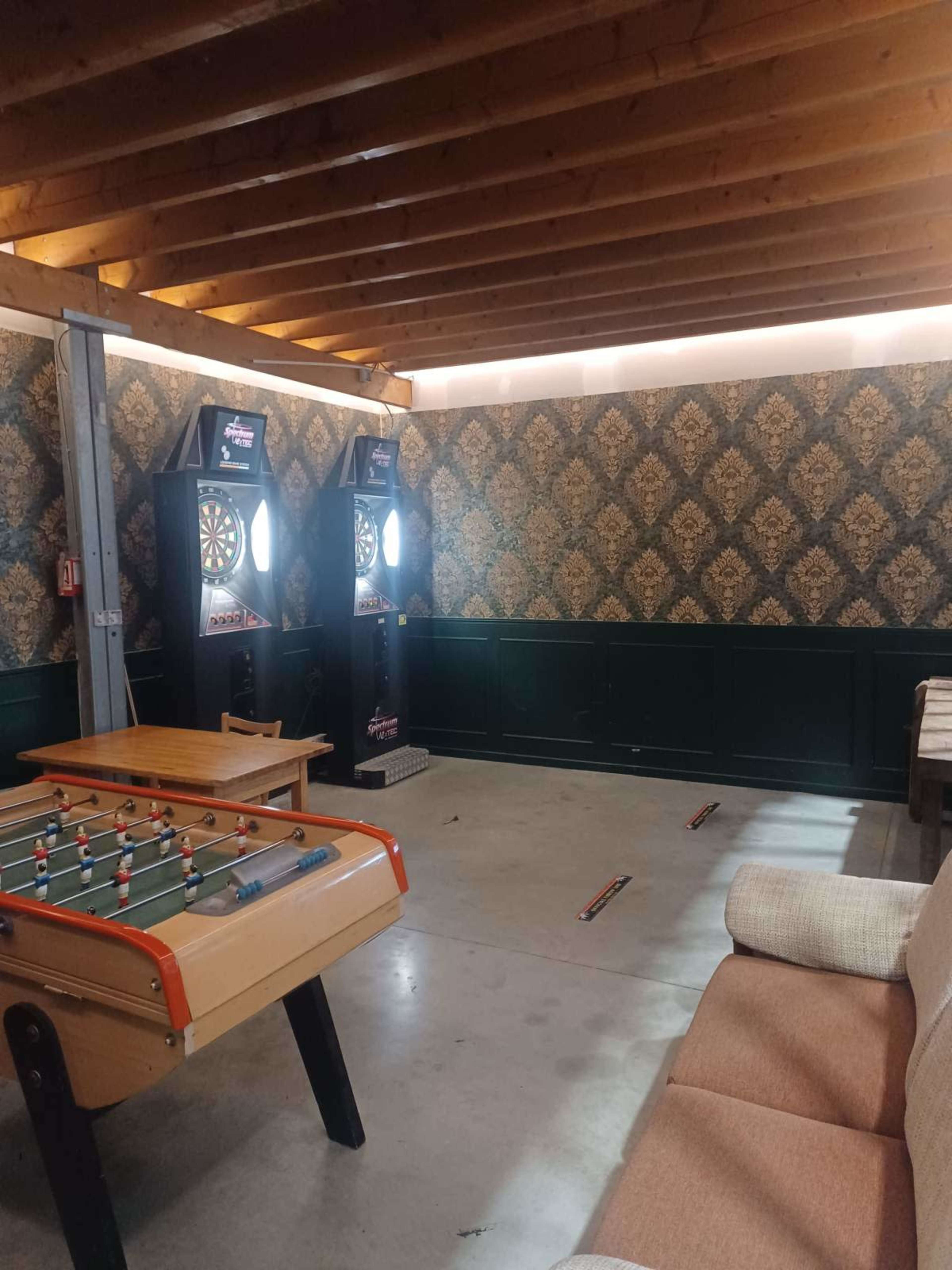 The scene shows a recreational room with dartboards, a foosball table, and a couch under a wooden ceiling.