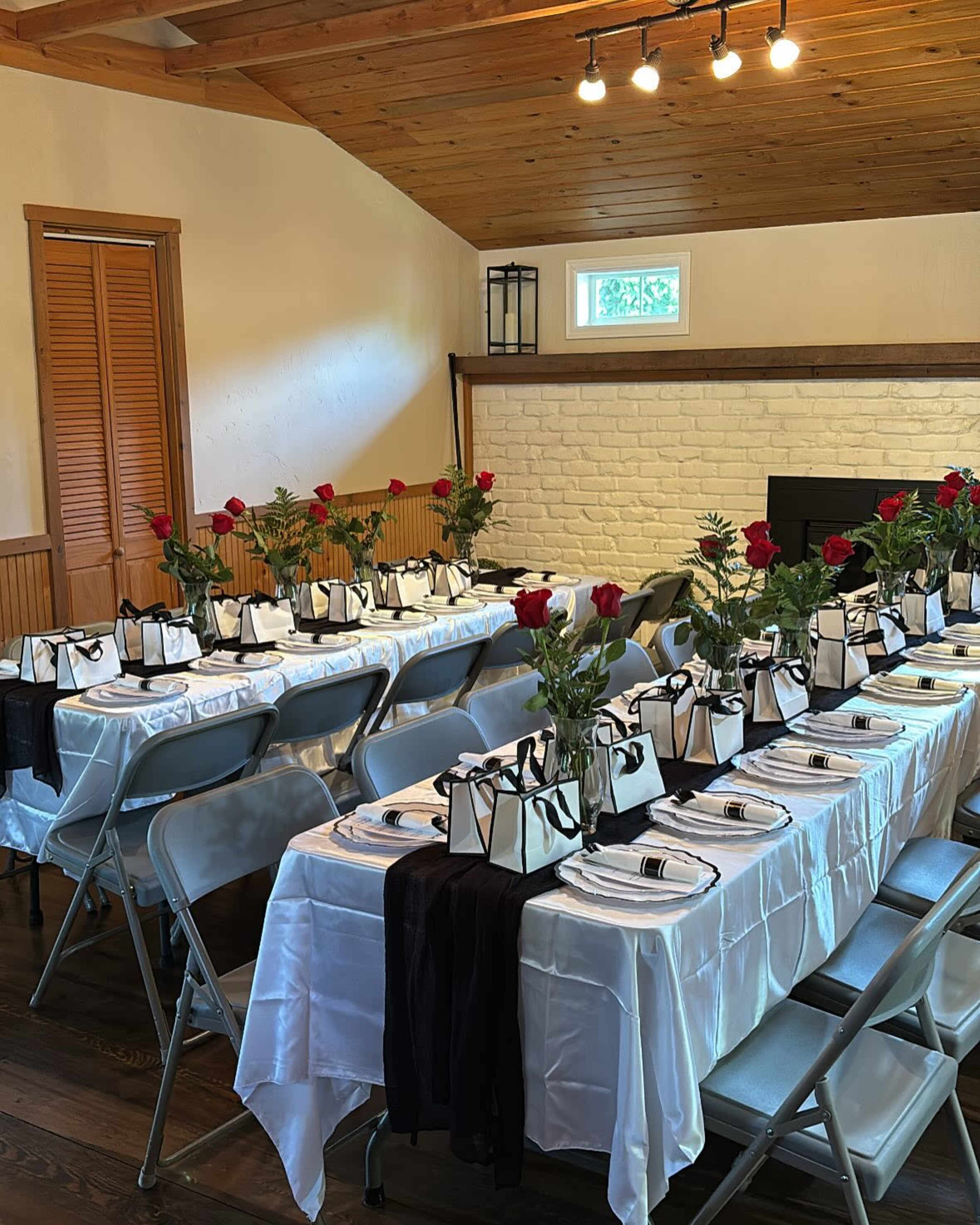 The image shows a dining setup with multiple tables covered in white tablecloths, each adorned with black and white bags and red rose centerpieces.
