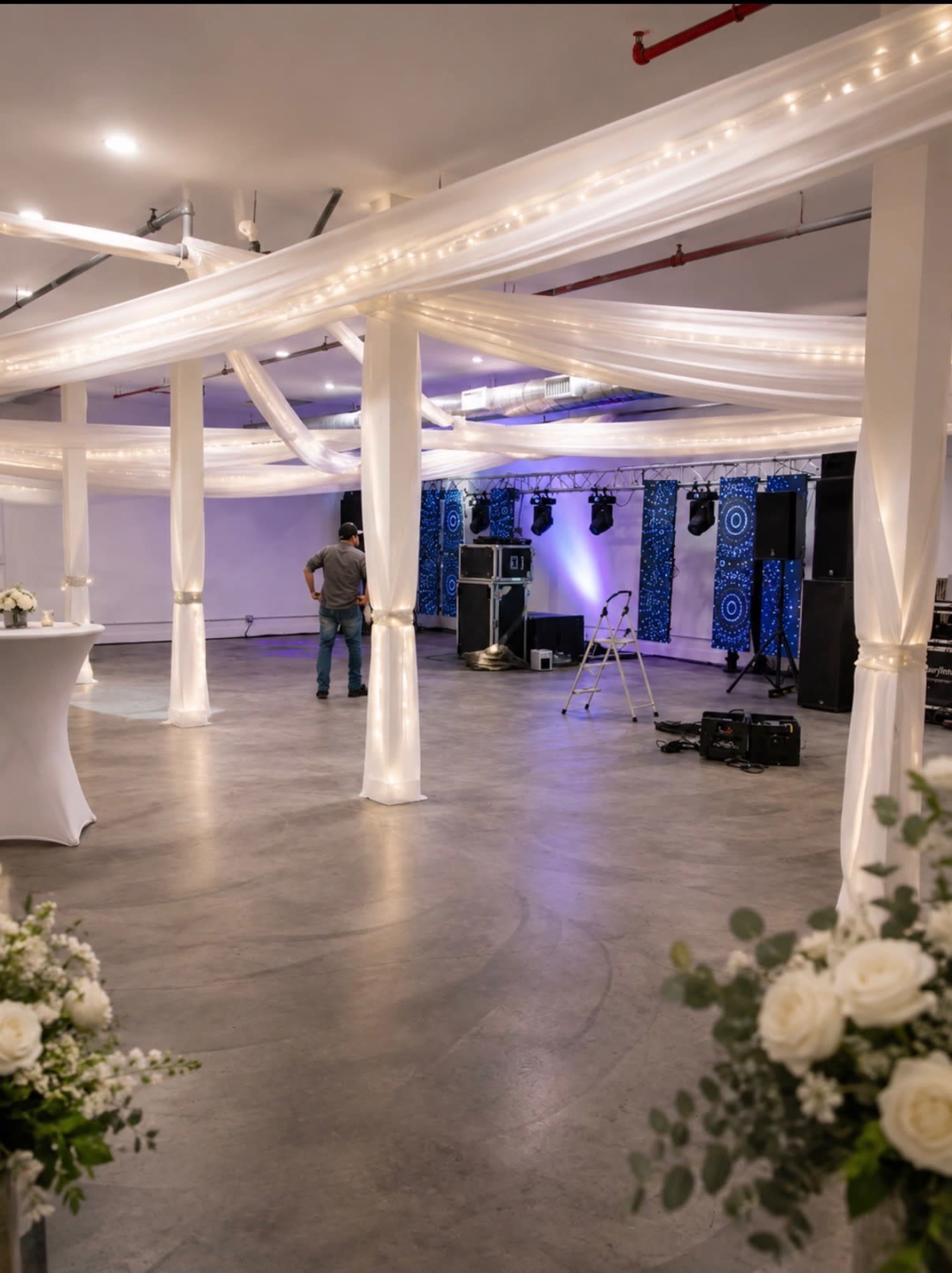 The image shows an empty event space with draped fabric and lighting, featuring a stage setup in the background and floral arrangements at the front.