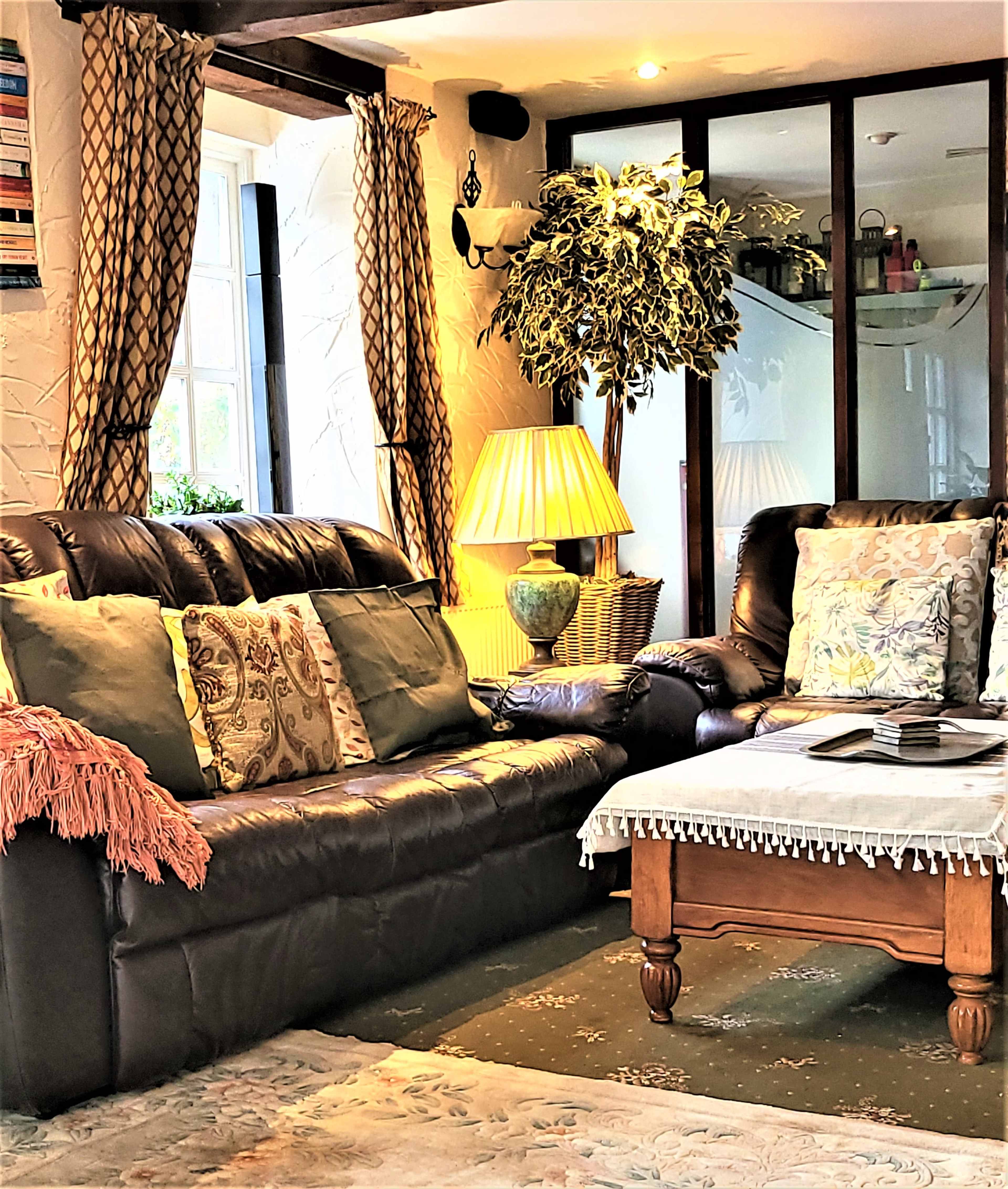 A cozy living room features dark leather sofas adorned with patterned cushions, a wooden table with a lace cloth, and a standing lamp beside a tall potted plant.