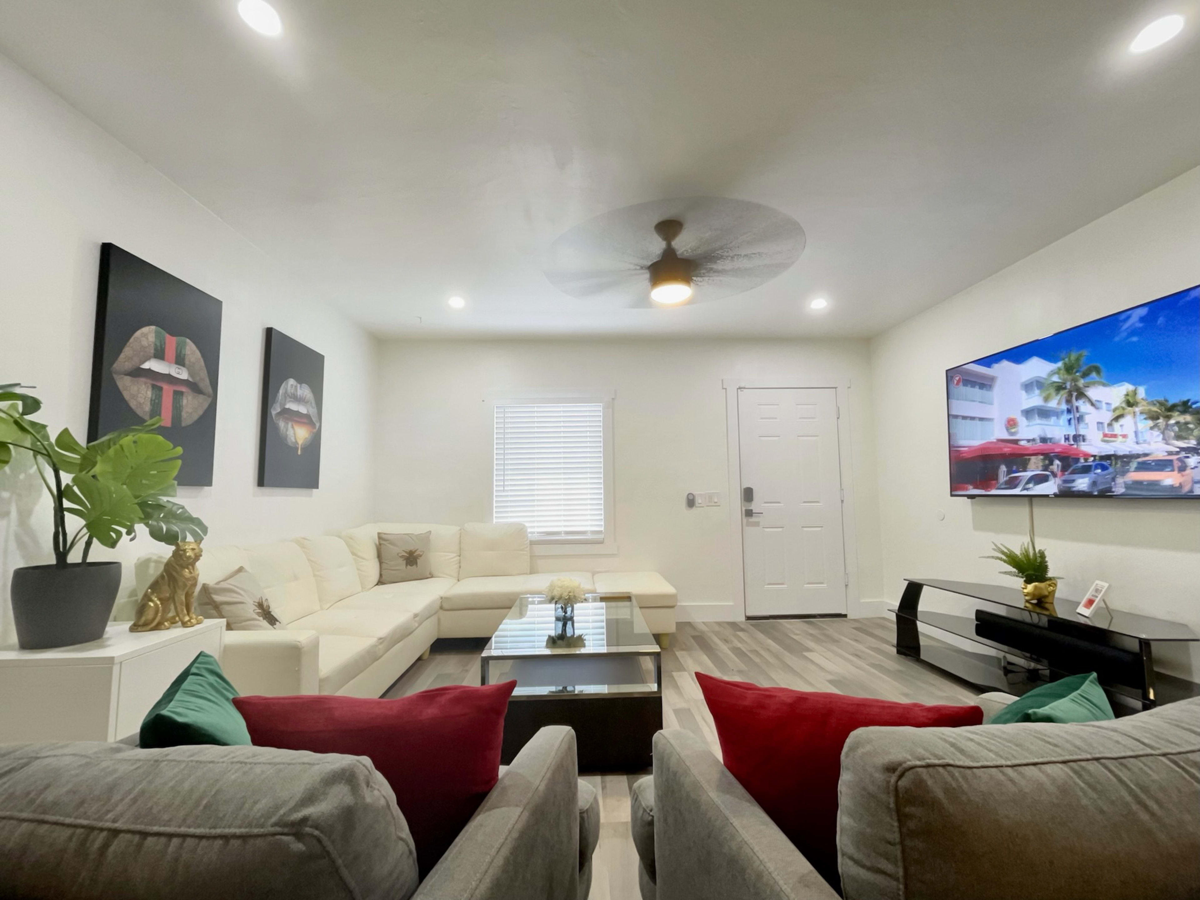 A modern living room with a white sectional sofa, two gray chairs with red and green cushions, a coffee table, wall art, and a large flat-screen TV.