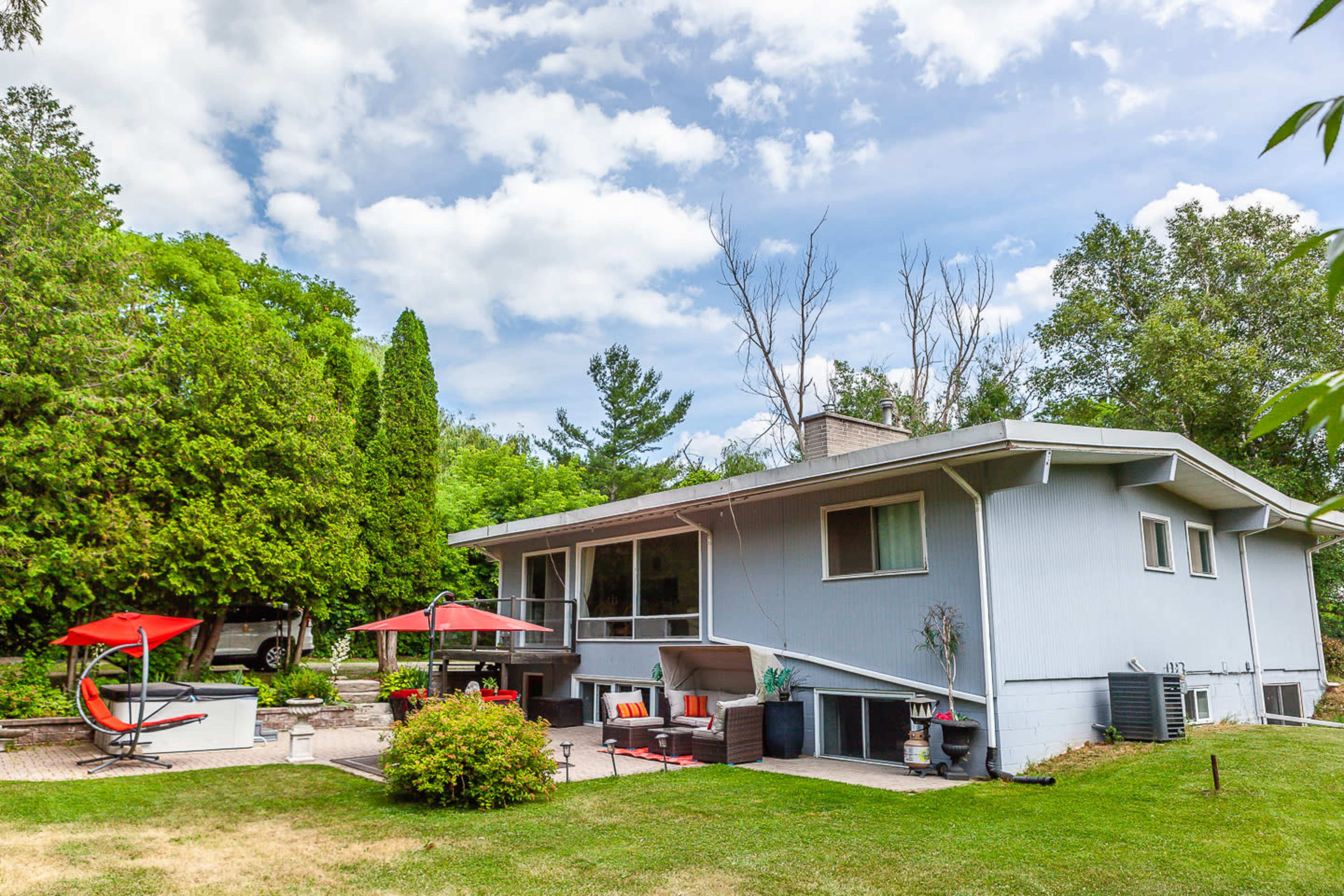 A light blue house with a landscaped yard features outdoor seating areas and a patio under red umbrellas.
