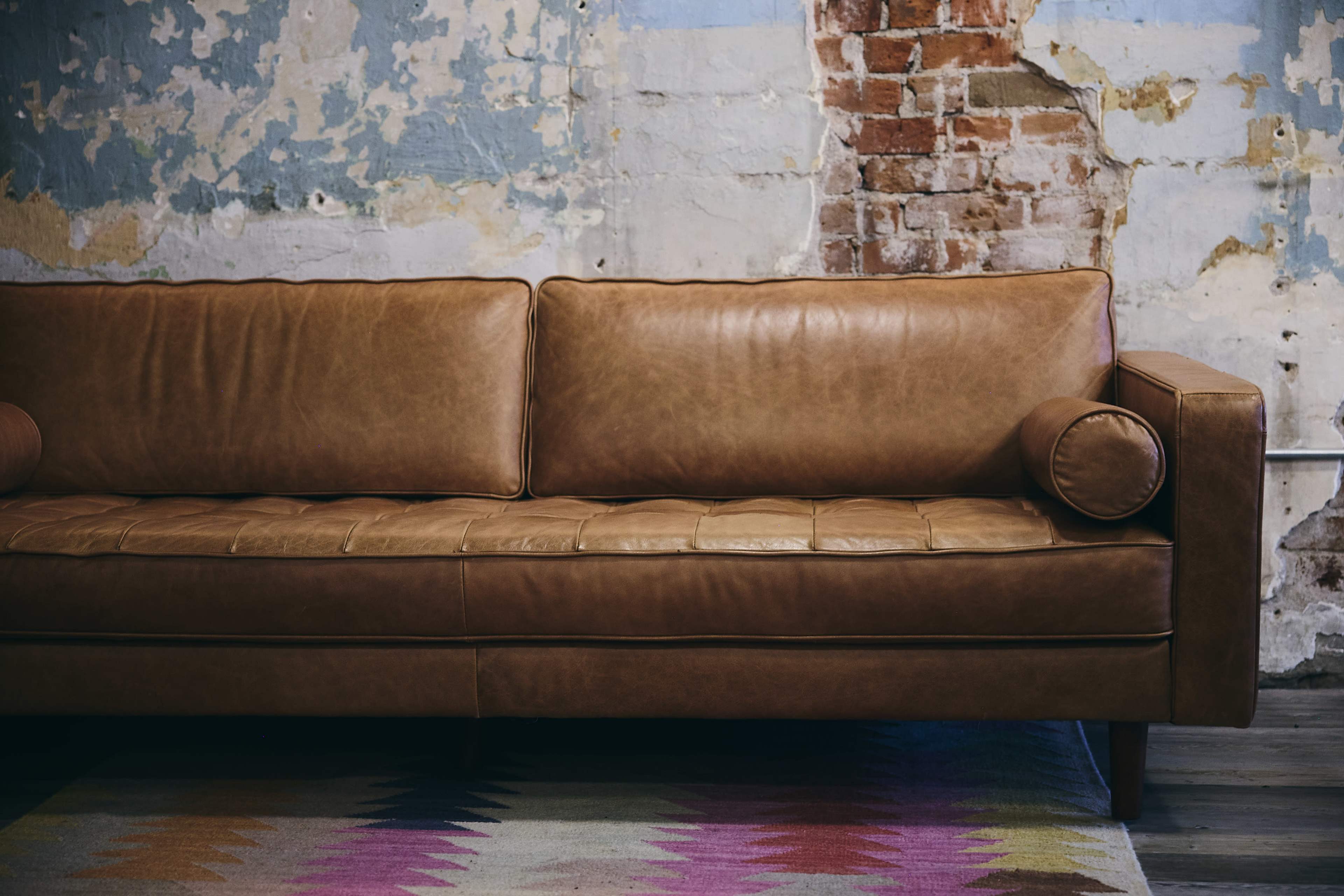A brown leather sofa with a round armrest is positioned against a wall featuring peeling paint and exposed brick.