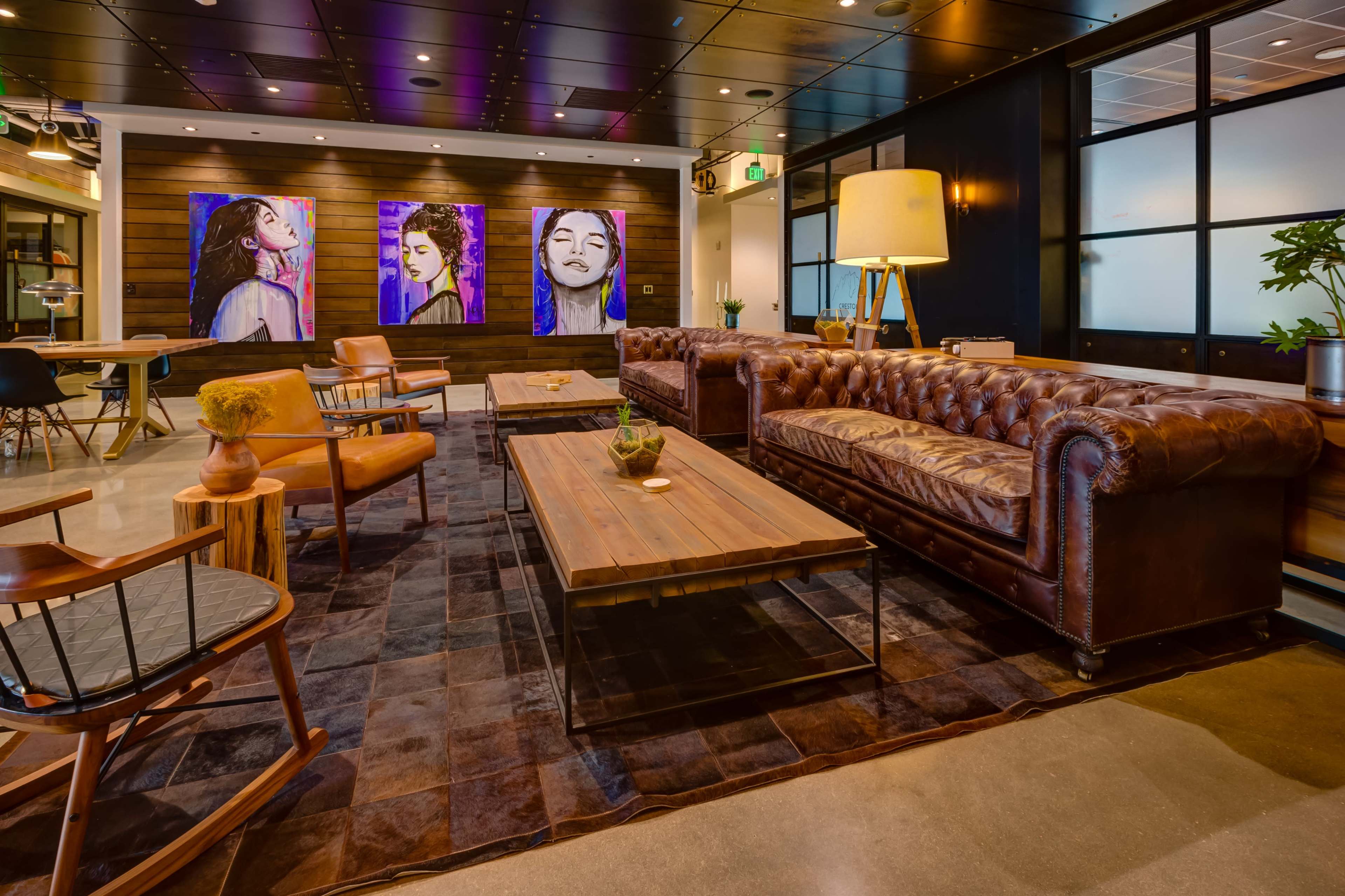 The image shows a modern lounge area featuring a leather couch, wooden tables, and colorful portrait artworks on the wall.