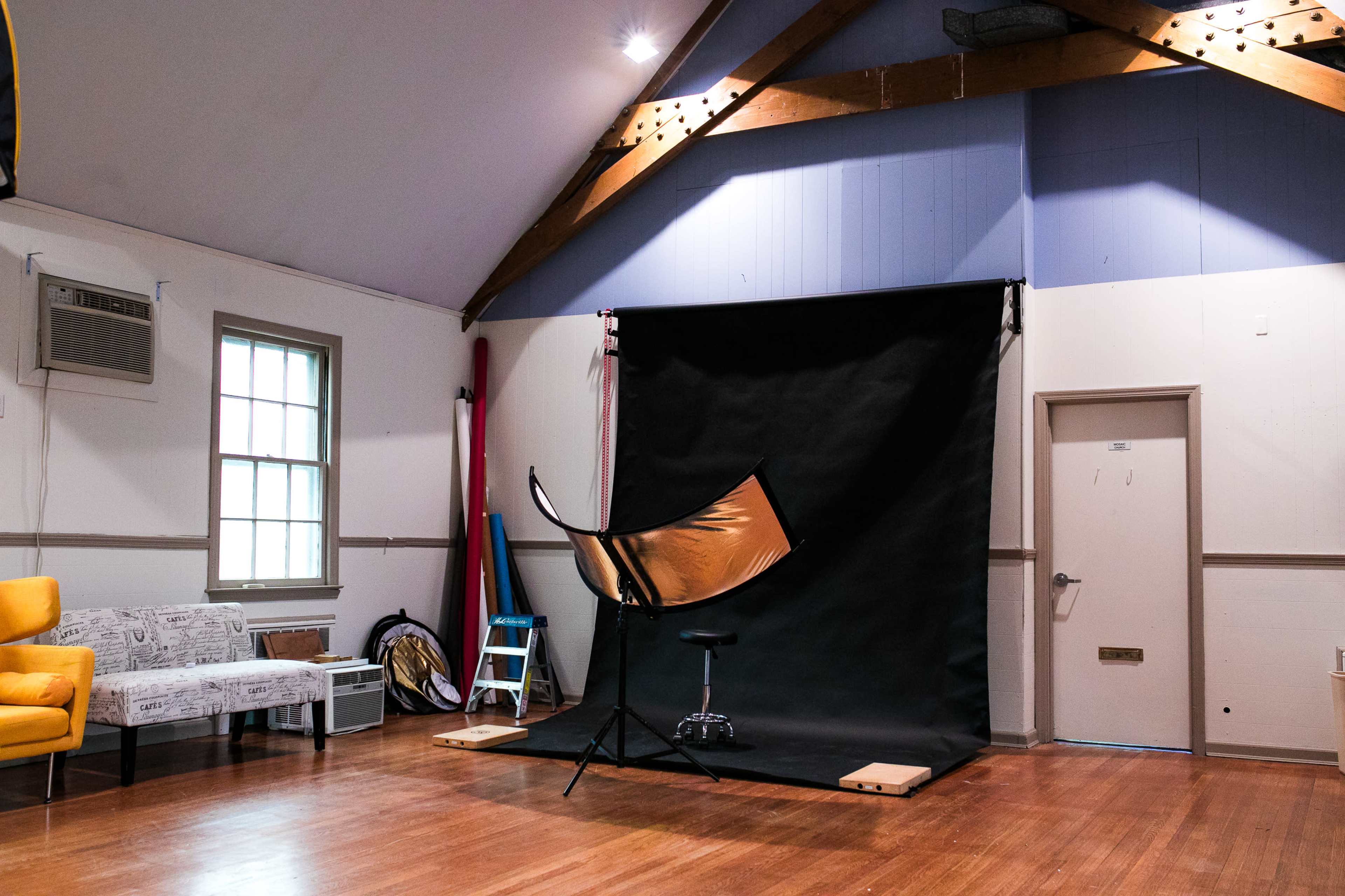 A spacious studio with a black backdrop, a stool, and a reflective umbrella on a stand, along with a couch and an air conditioning unit.