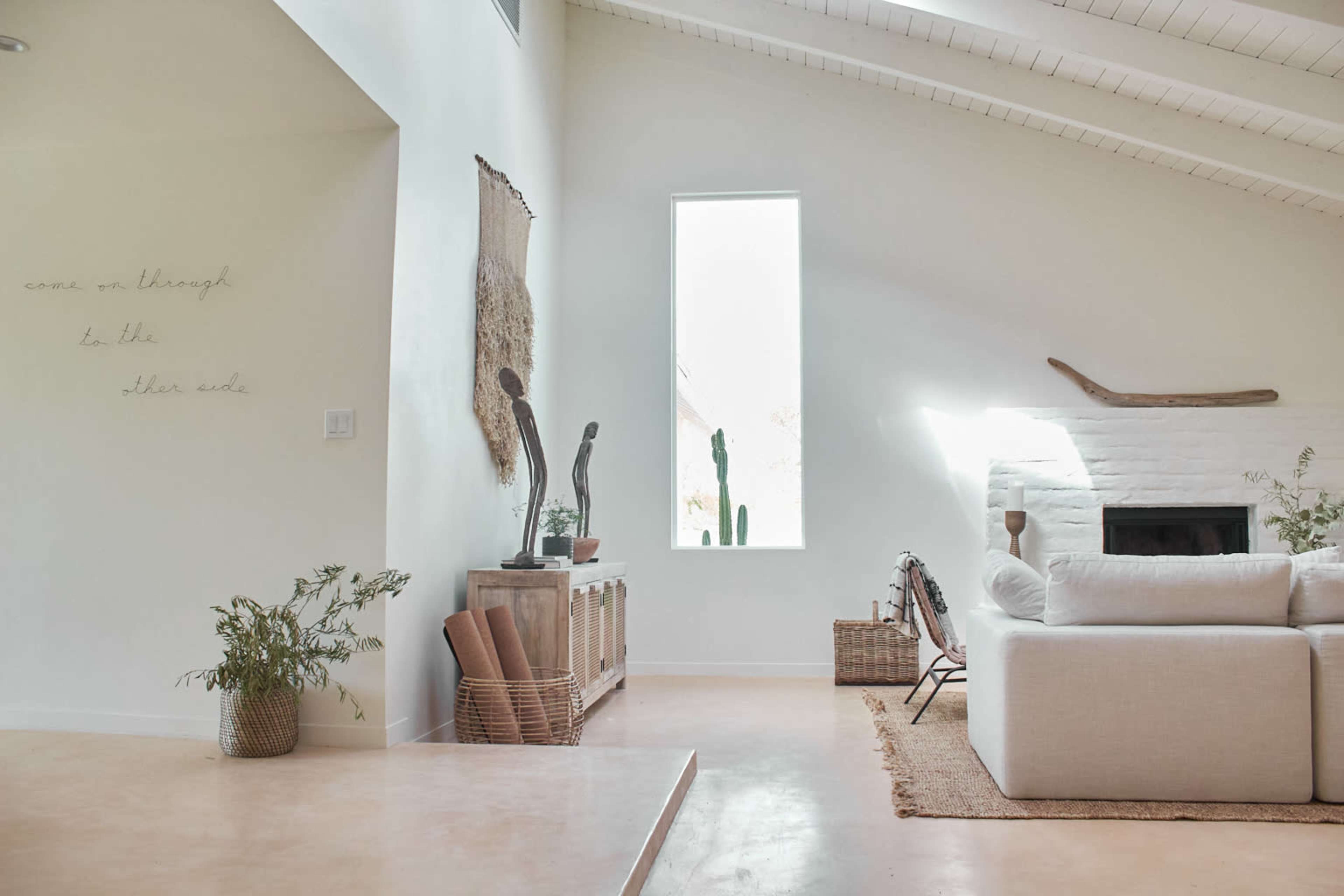 The image shows a modern, minimalist living room with a white sofa, a light-colored floor, and large windows that allow natural light to enter, featuring decorative elements and plants.
