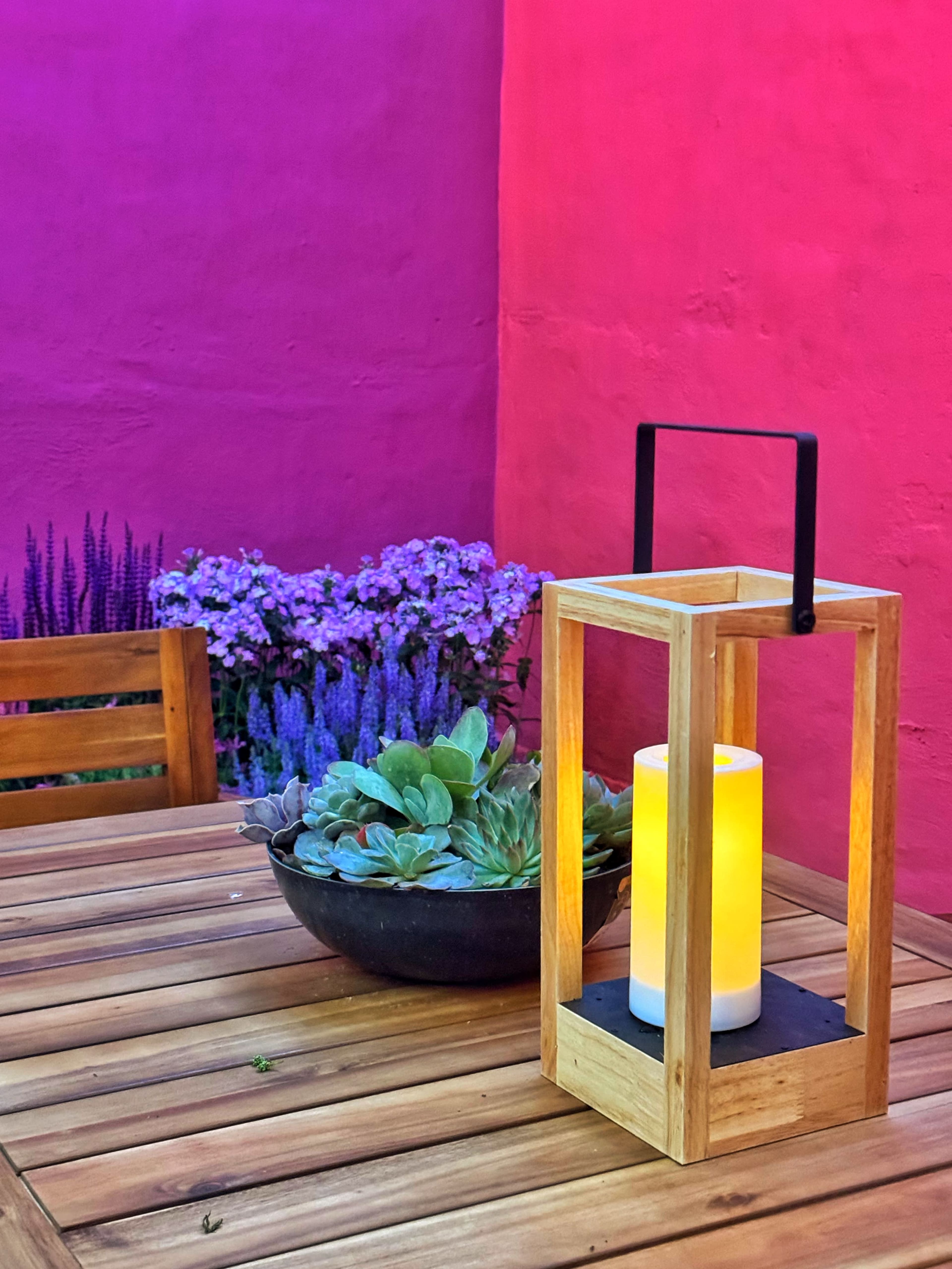 A wooden lantern with a candle is placed on a tabletop beside a bowl of succulents, with a vibrant purple wall and flowers in the background.