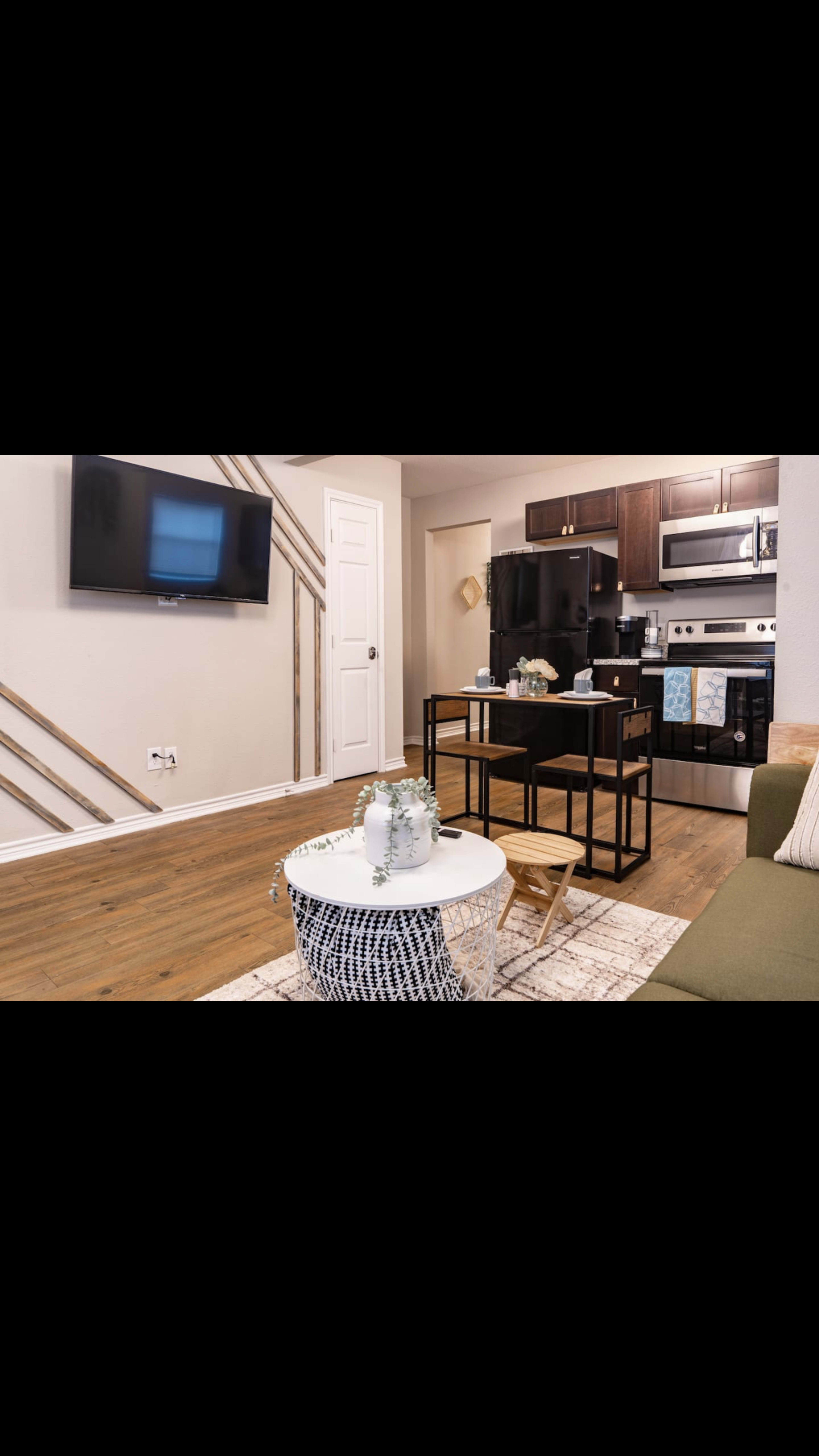 A modern living room area with a green sofa, a low coffee table, and a small dining table, adjacent to a kitchenette featuring a black refrigerator and oven.
