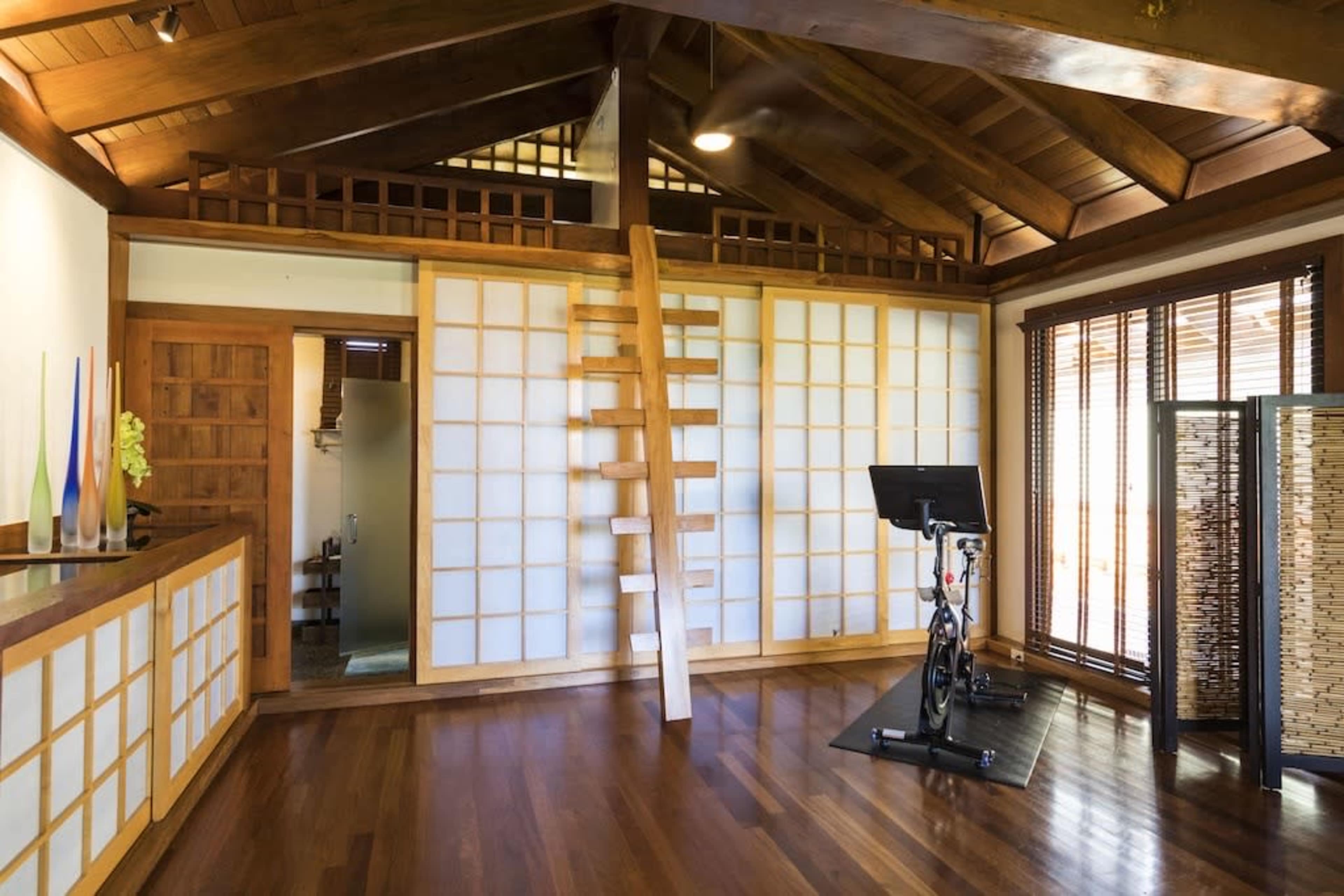 A wooden interior space featuring a loft area with a ladder, sliding shoji-style doors, and a stationary exercise bike on a polished floor.