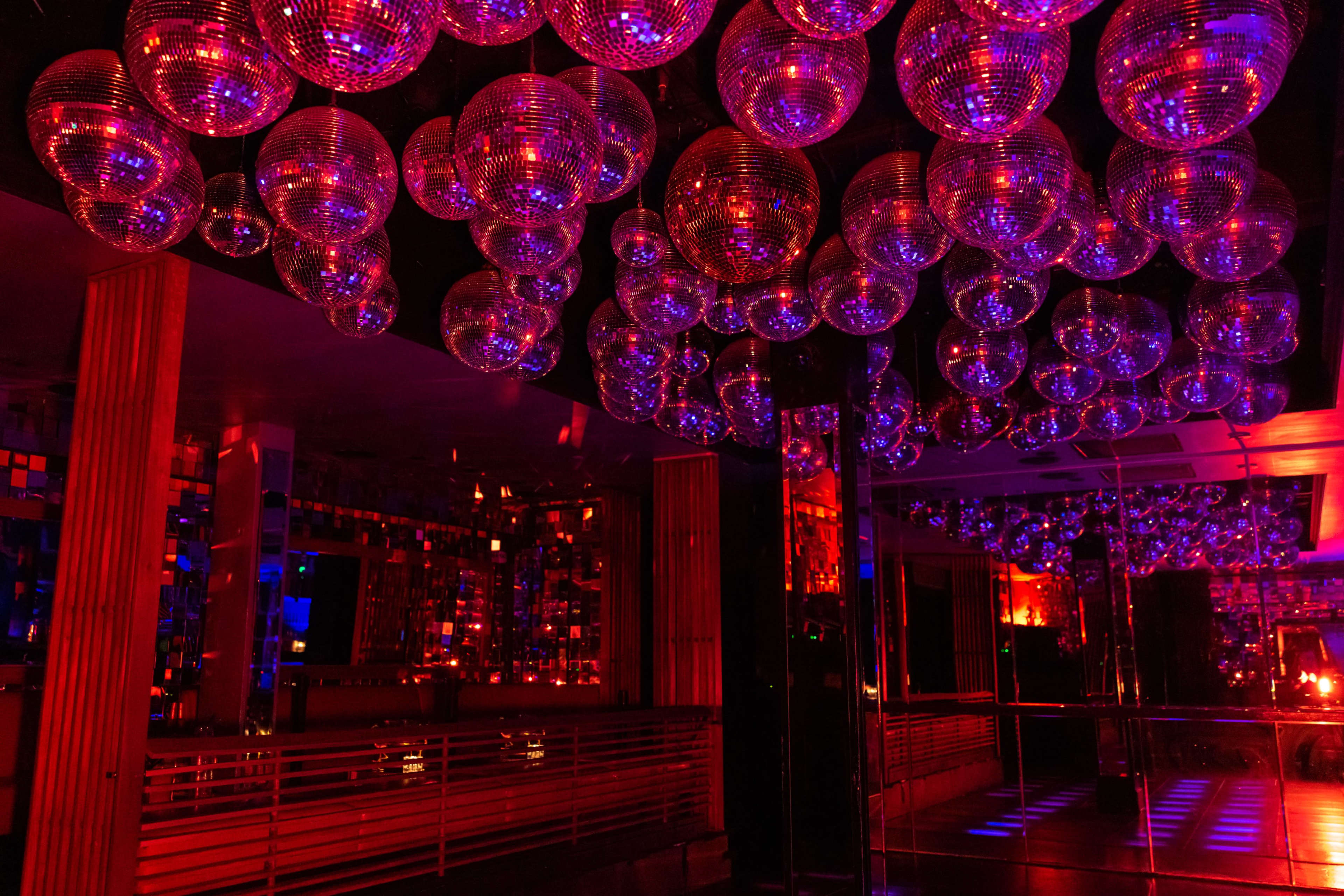 A nightclub interior with multiple reflective disco balls hanging from the ceiling, illuminated by red and blue lights.