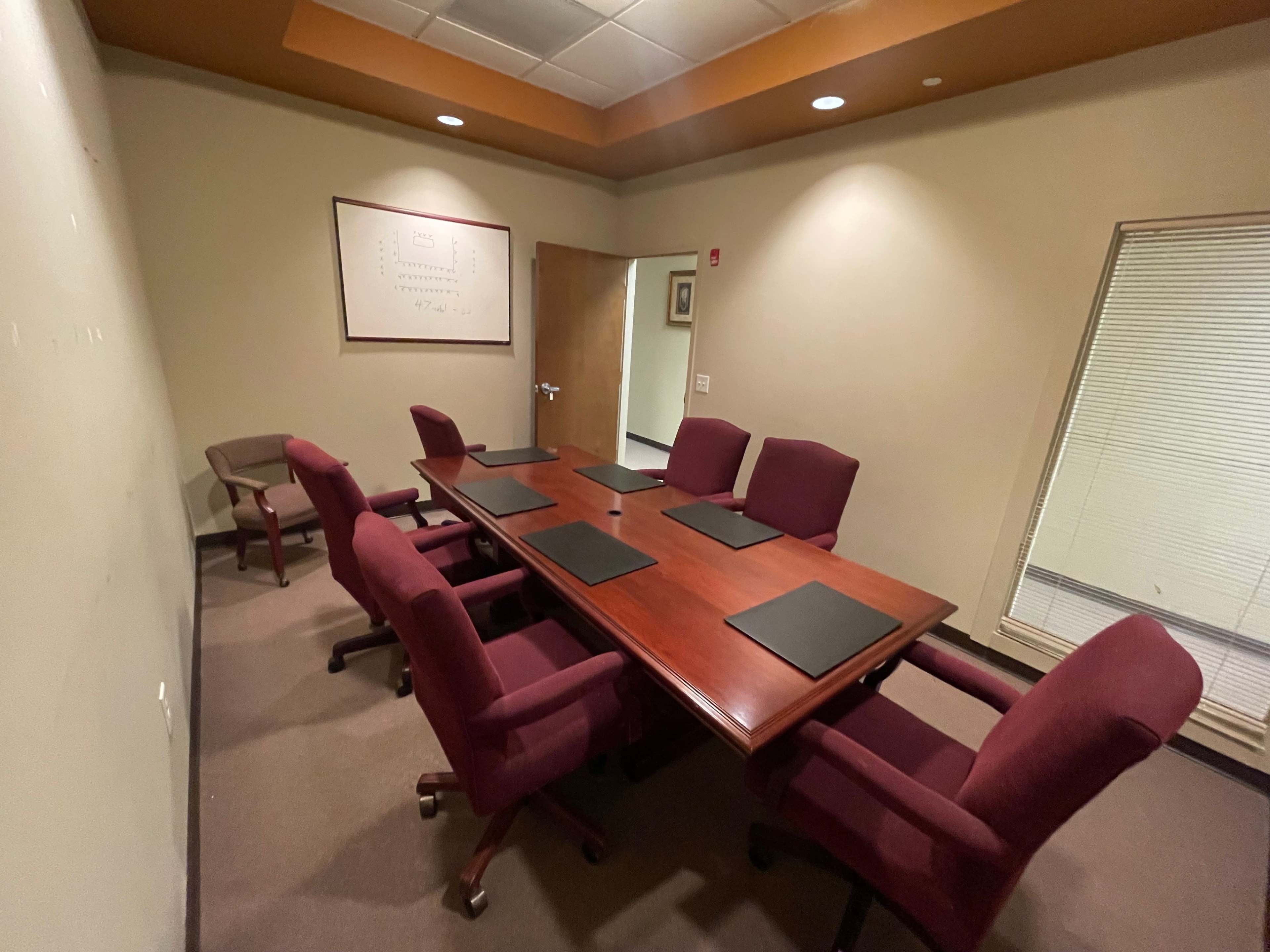 A conference room features a large wooden table surrounded by six maroon chairs, with a whiteboard and a closed door visible in the background.