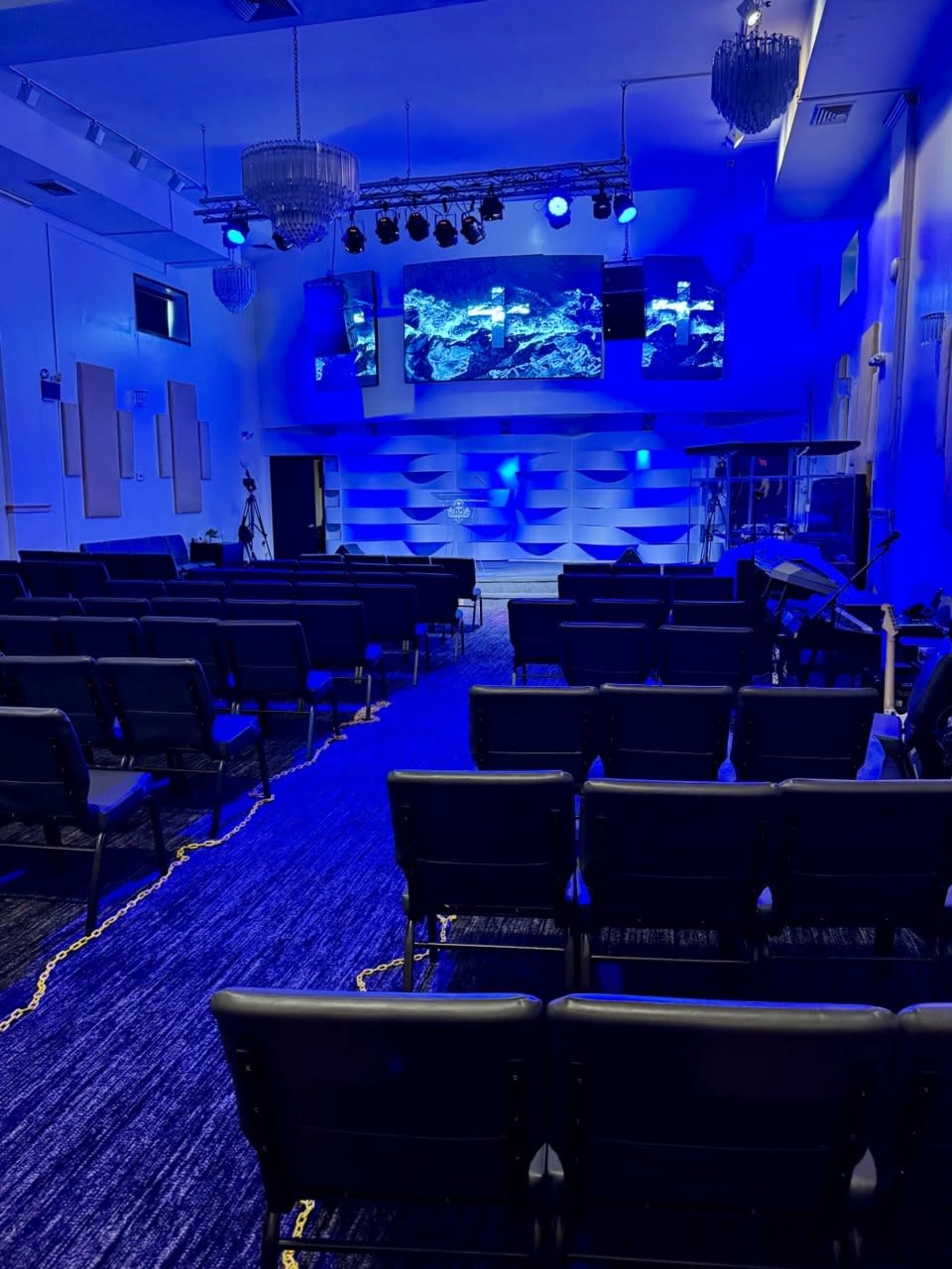 The image shows an empty auditorium with rows of black chairs, illuminated by blue lighting and featuring a stage with large screens displaying visuals.