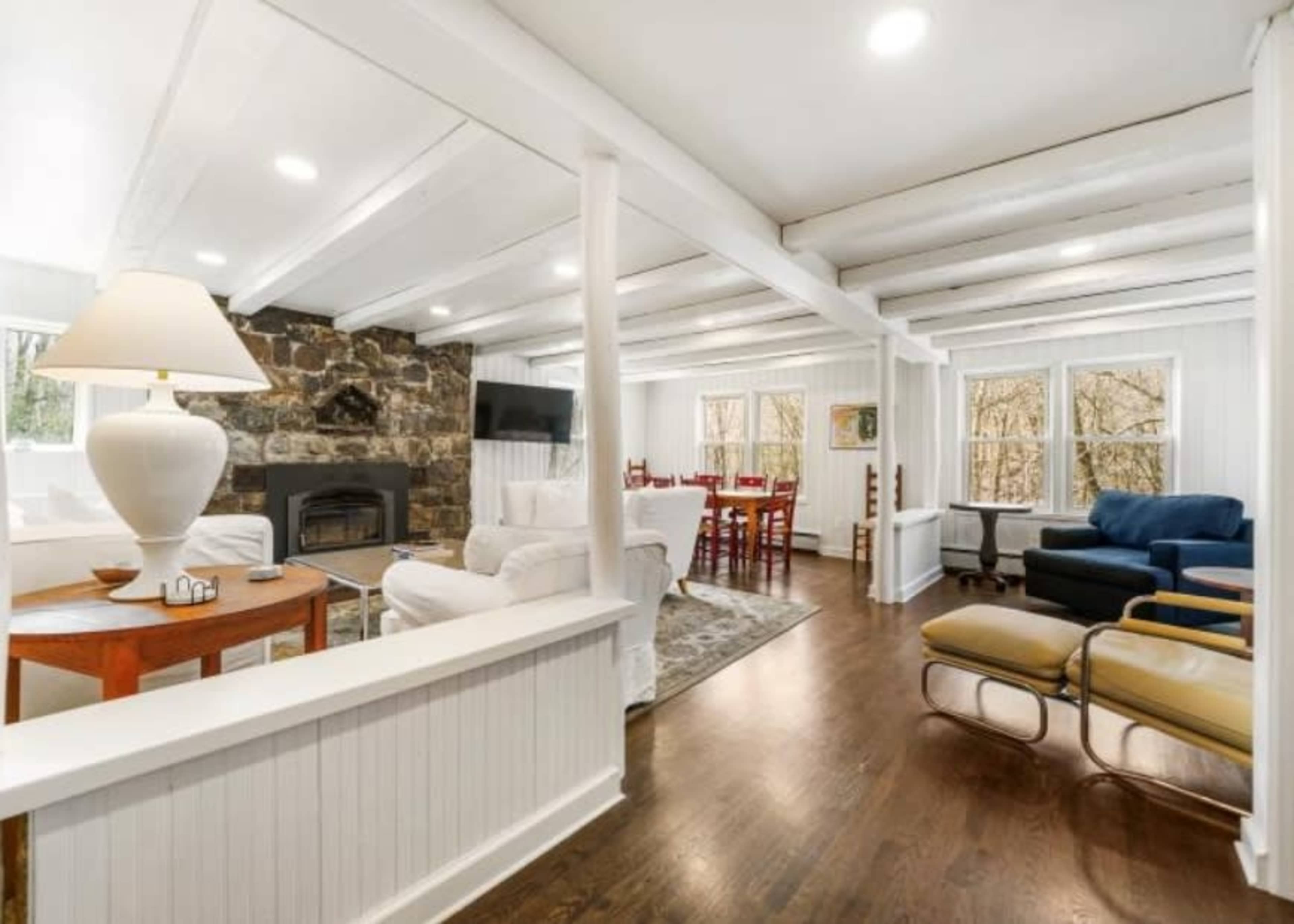 The image shows a cozy living area with a stone fireplace, white walls, and wooden beams, featuring a mix of seating options and a dining space in the background.