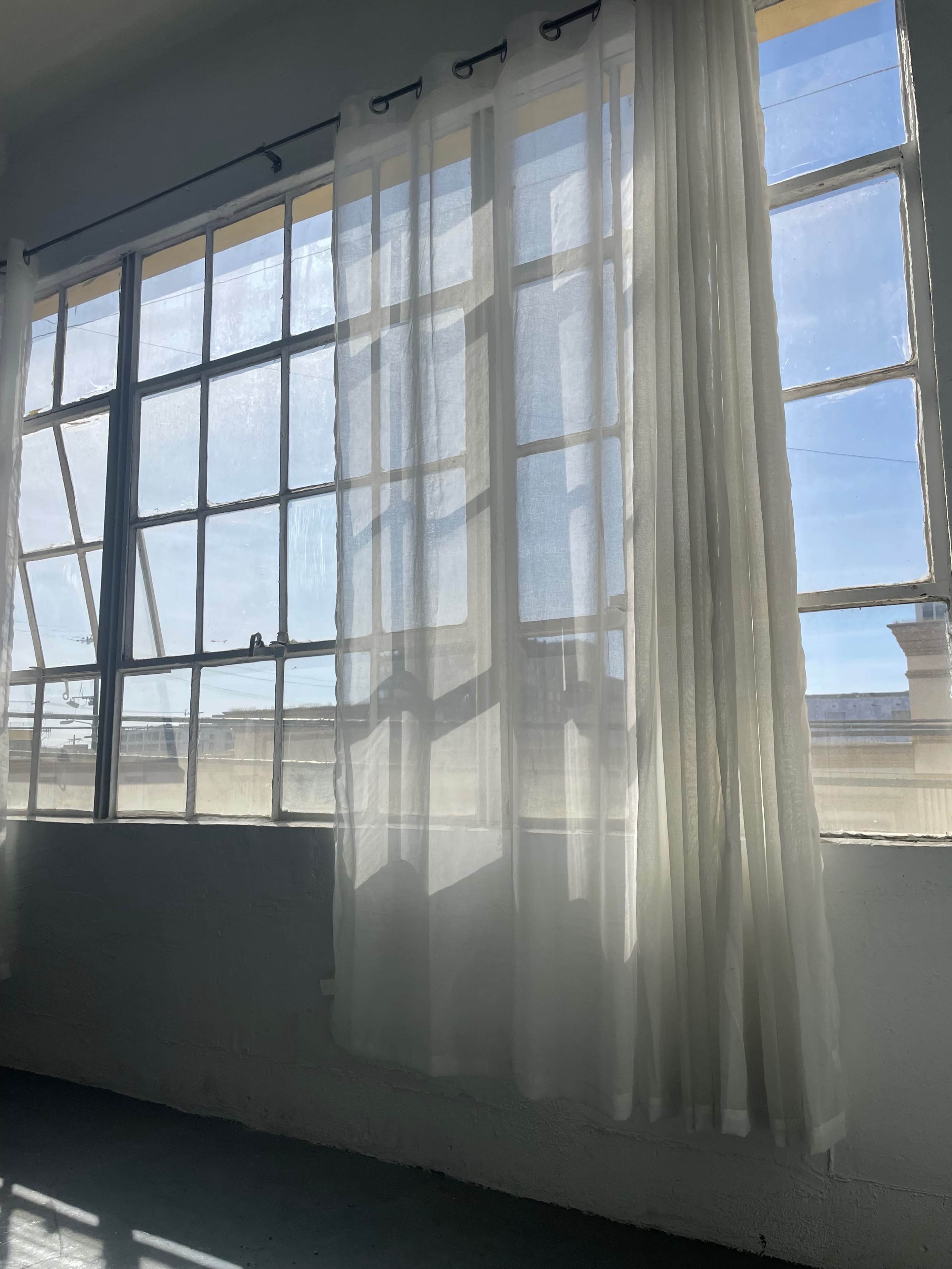 The image shows sunlight filtering through sheer curtains in front of large windows in a minimalist interior space.