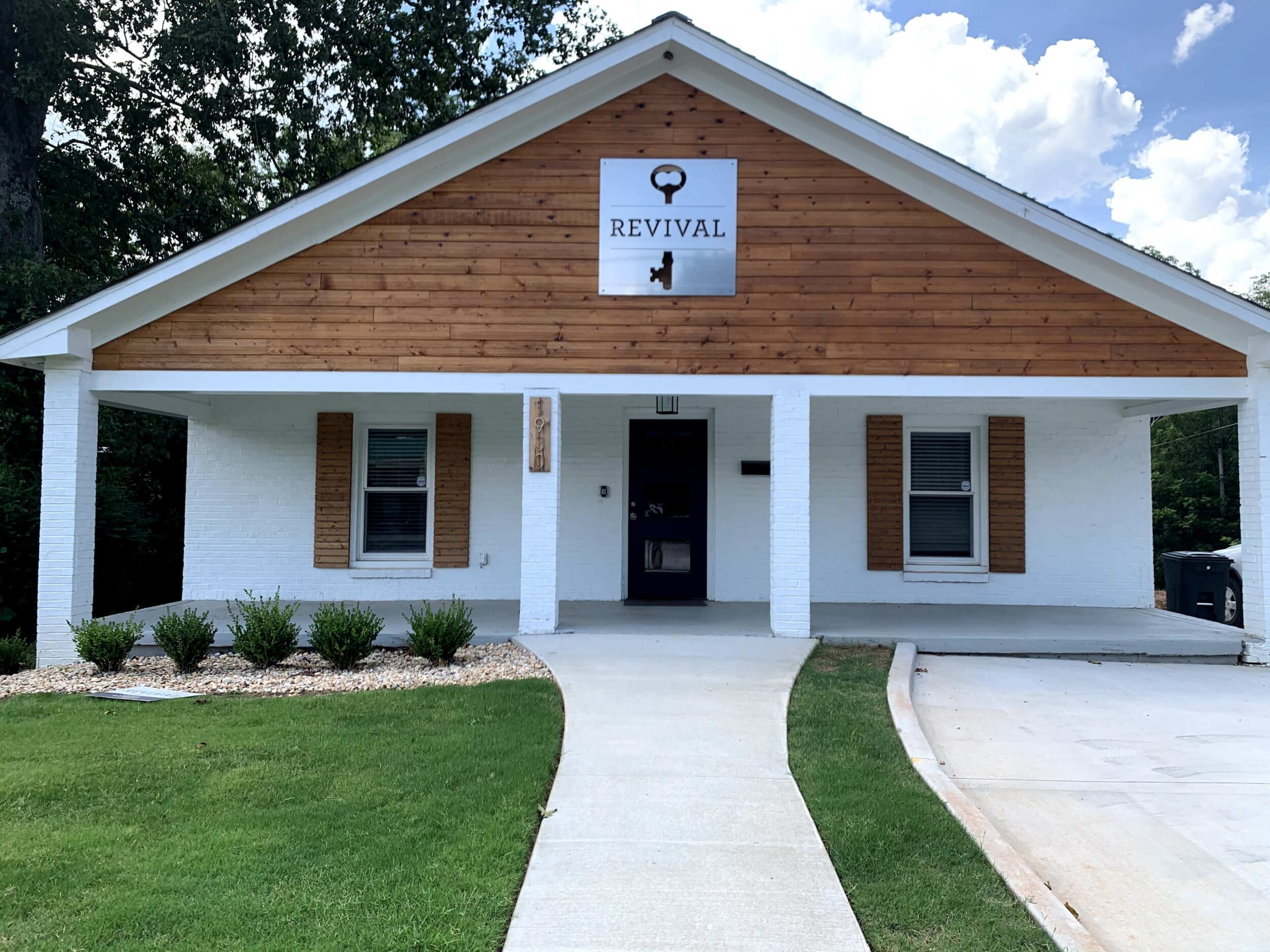 The image shows a single-story building with a wooden facade, featuring a sign that reads "REVIVAL" above the entrance and surrounded by a landscaped area.