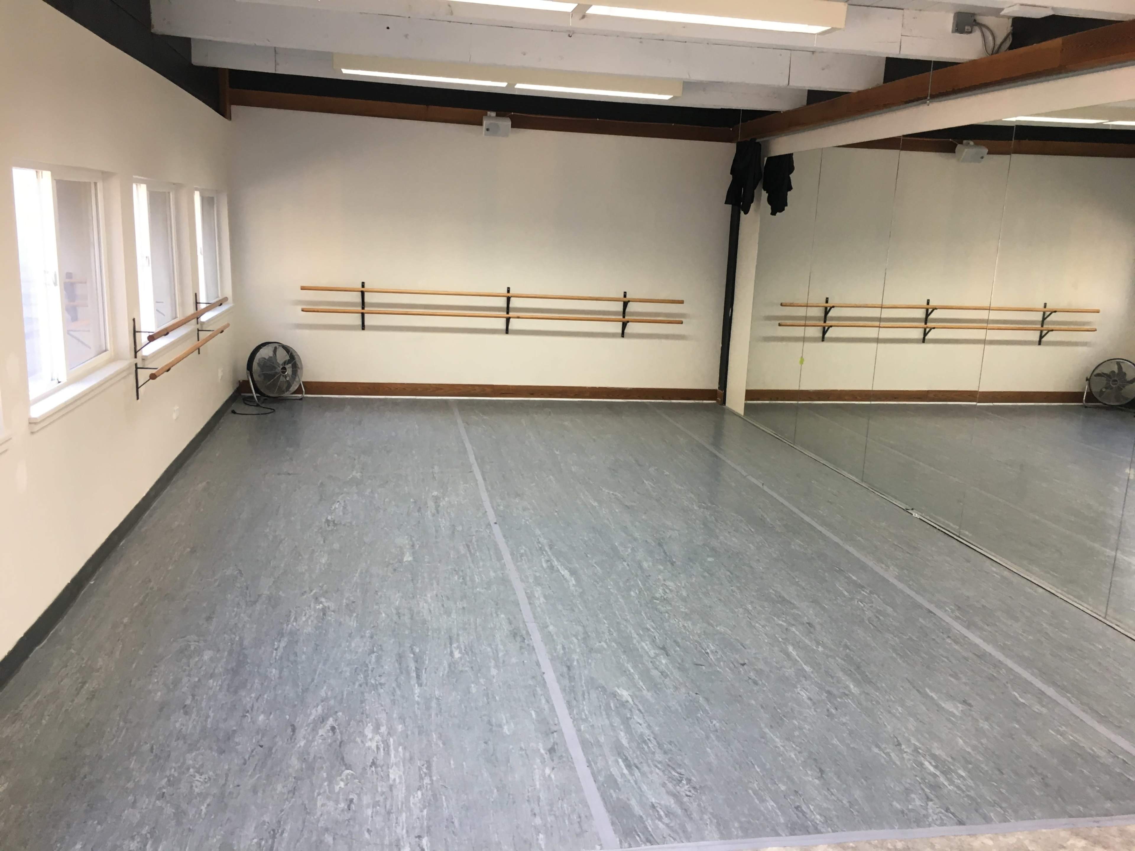 A mirror-walled dance studio with a wooden barre mounted along the wall and a fan in the corner.