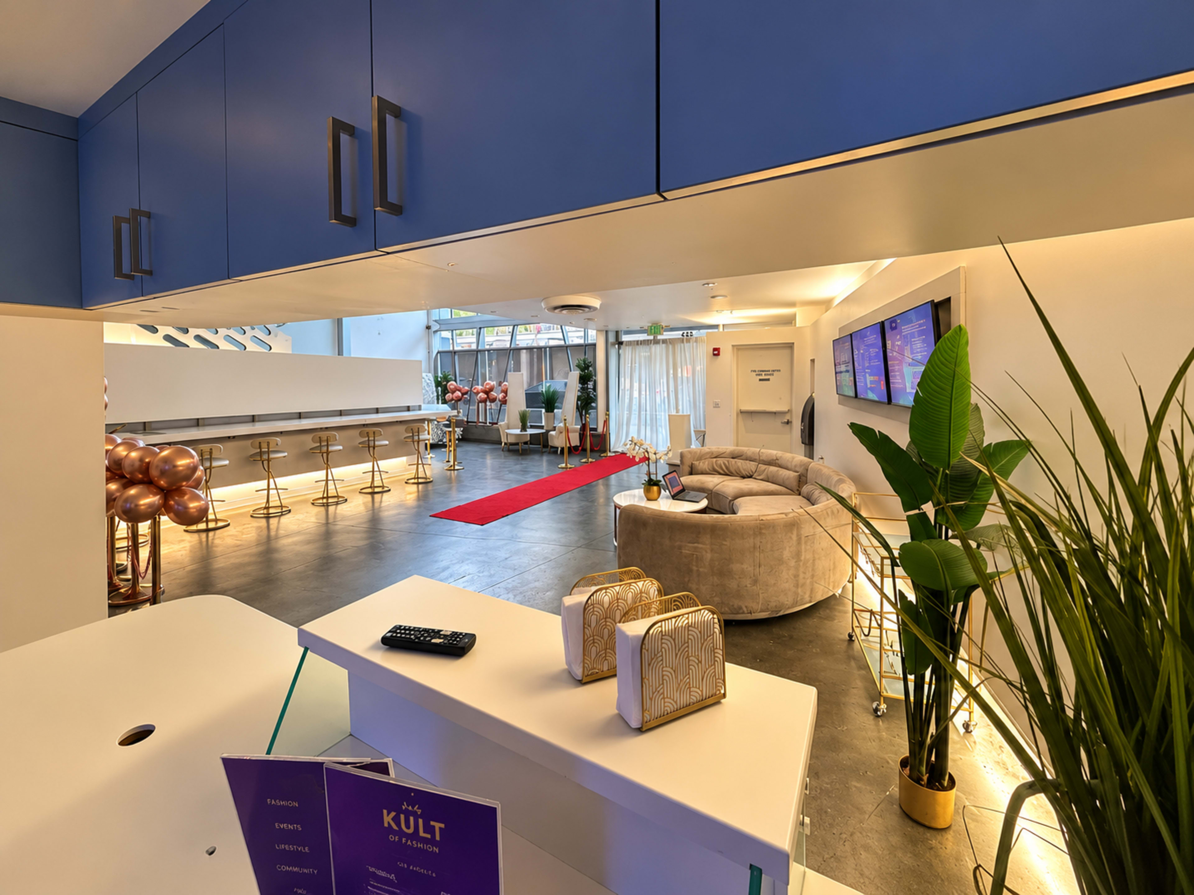 The image shows a modern reception area with a curved gray sofa, a long bar counter, and a red carpet leading to a spacious lounge decorated with balloons and plants.