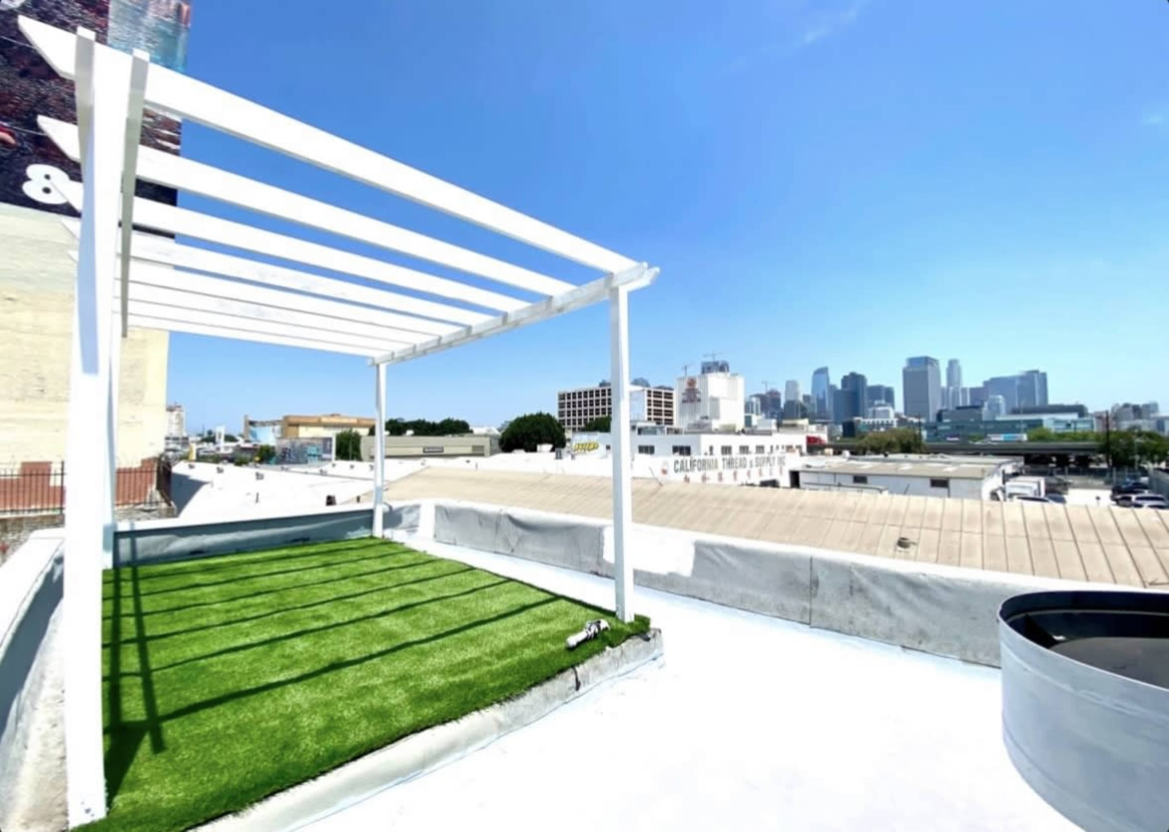 The image shows a rooftop area with artificial grass, a white trellis structure, and a skyline view of a city in the background.