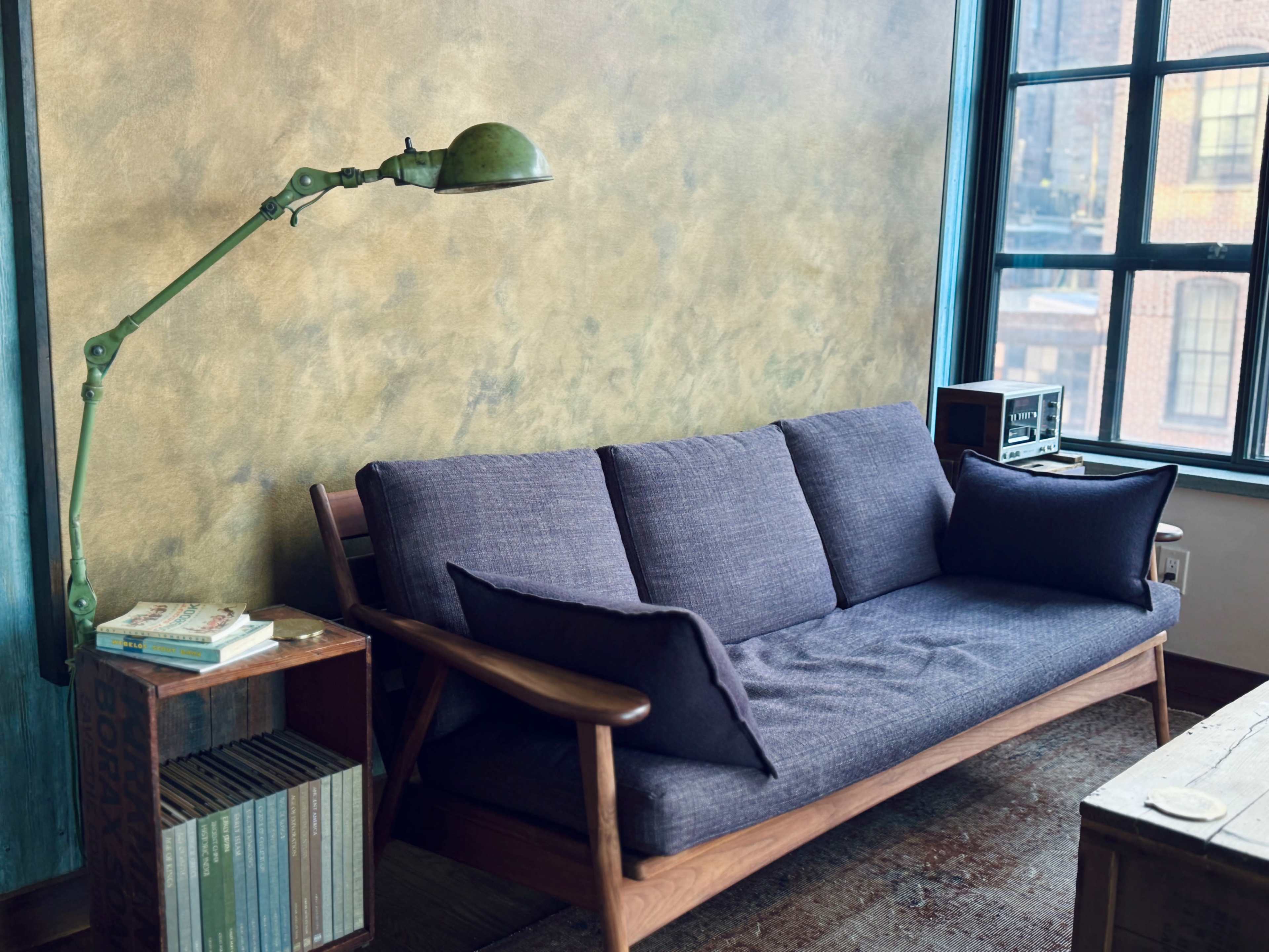 The image shows a modern interior with a dark blue sofa, a green arc lamp, a wooden side table with books, and a window allowing natural light to enter the room.