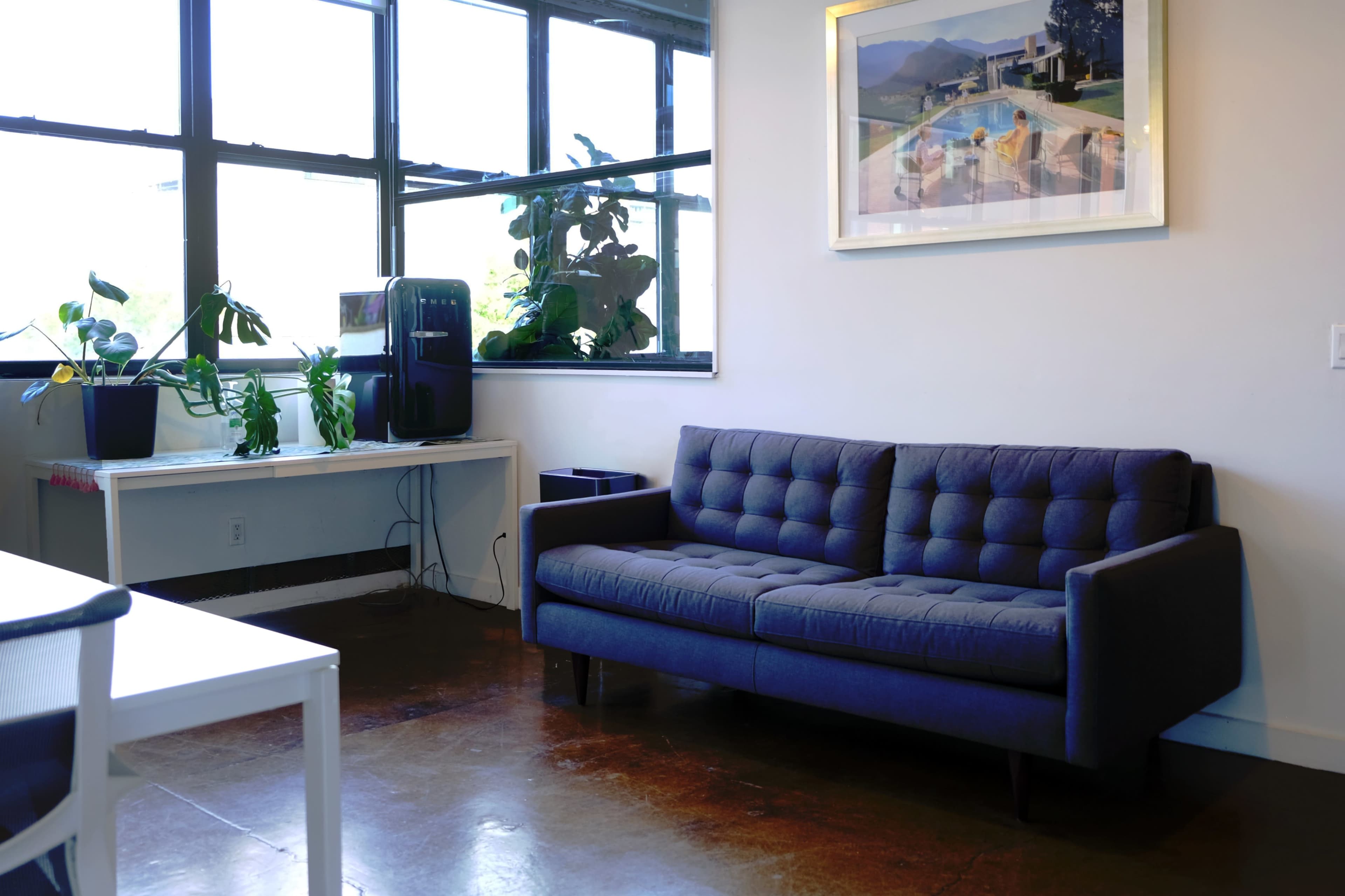 The image shows a modern living room with a blue couch, a white table, and large windows allowing natural light, alongside potted plants and a water cooler.