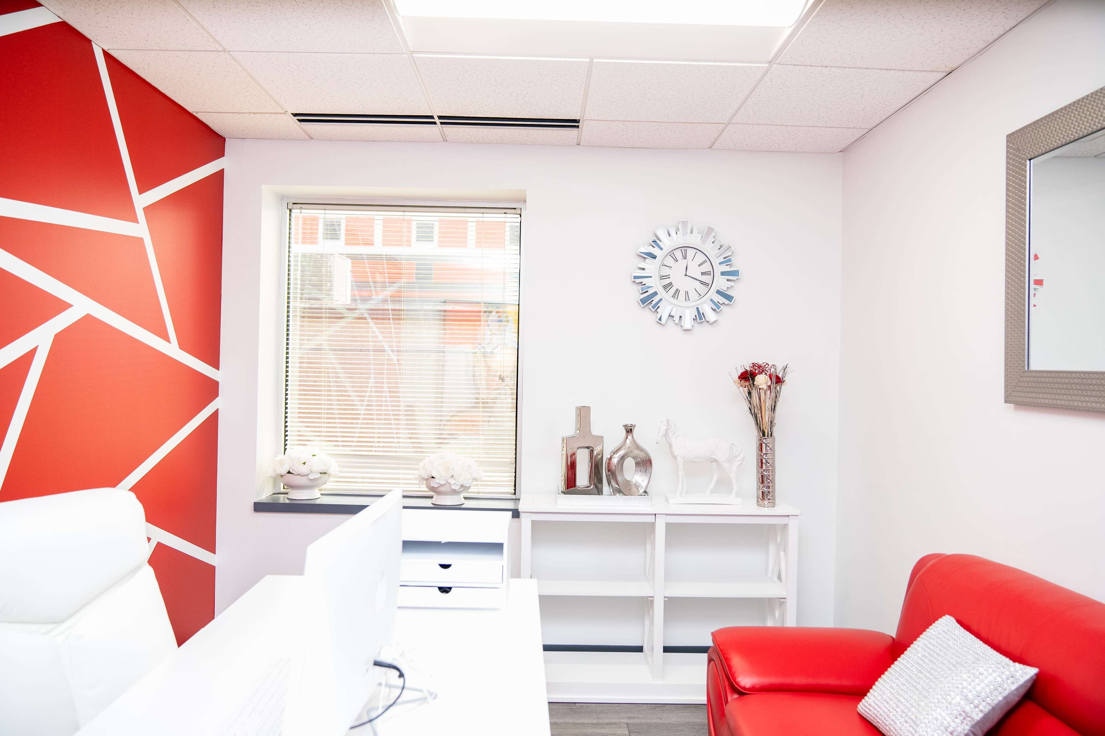 A modern office space with a red accent wall, a white desk with a computer, and a red couch beside a decorative shelf.