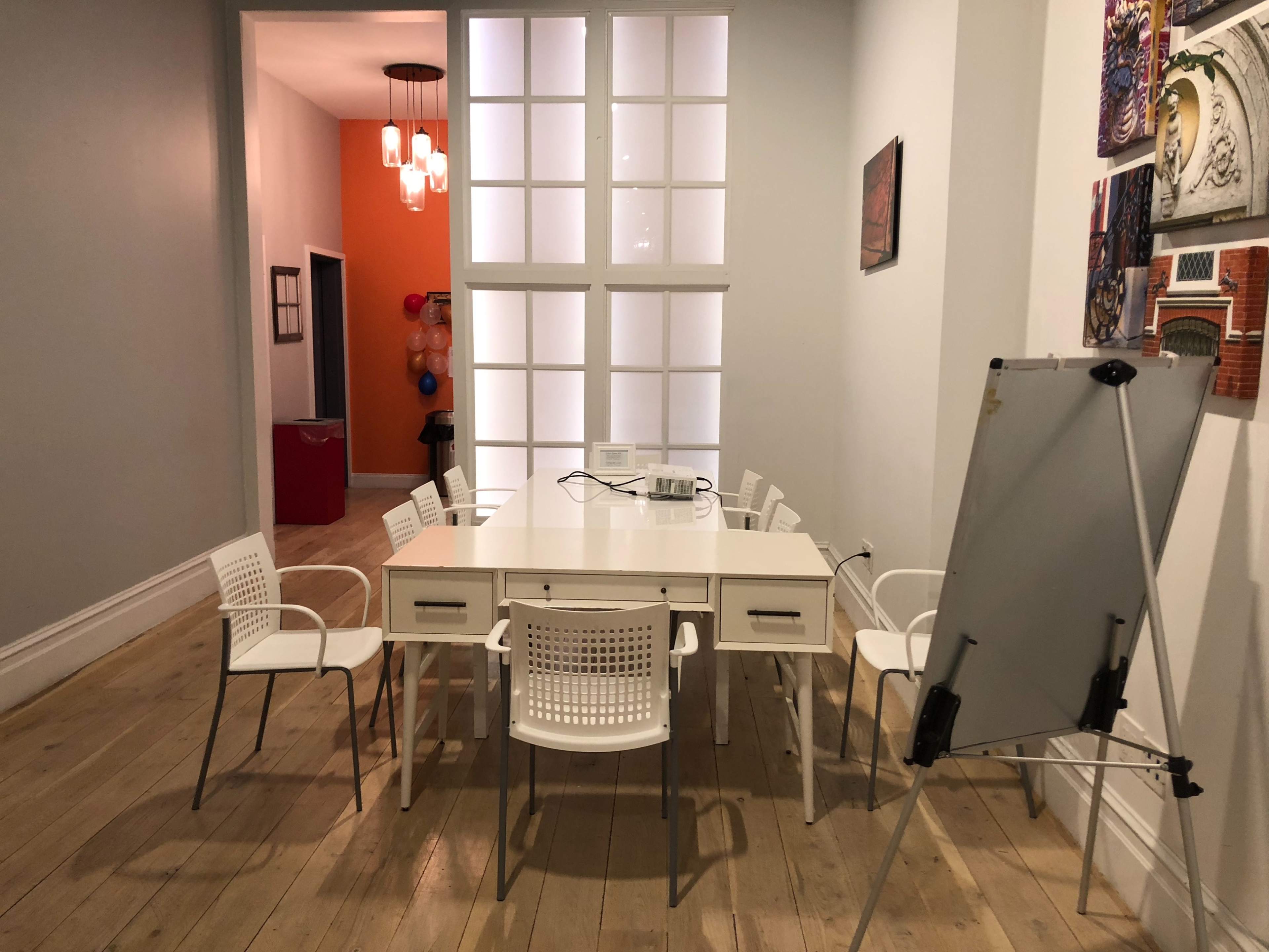 A bright meeting room features a long white table surrounded by white chairs, with a whiteboard at one end and a door leading to an orange-walled corridor.