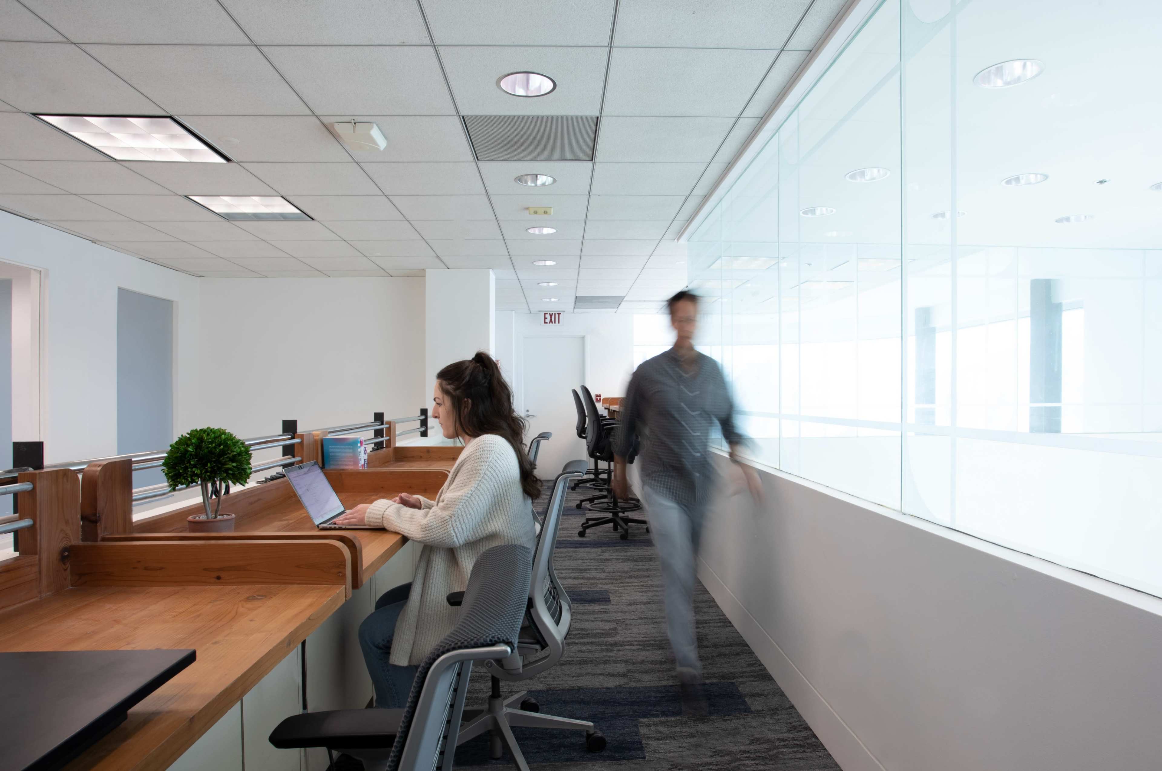 A woman sits at a wooden desk using a laptop while a man walks past a glass wall in a modern office space.