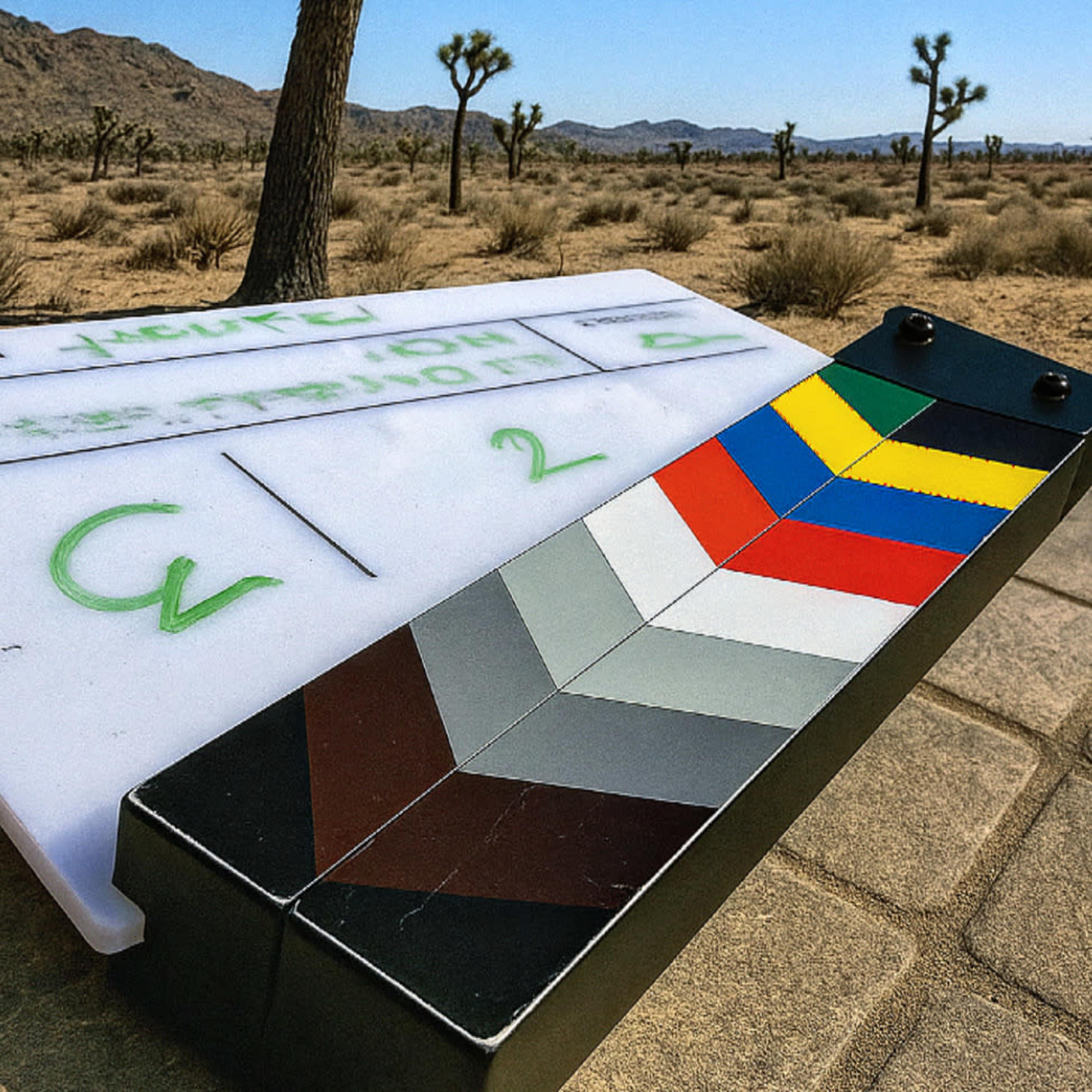 A film clapperboard rests on a stone surface in a desert landscape with Joshua trees and distant mountains.