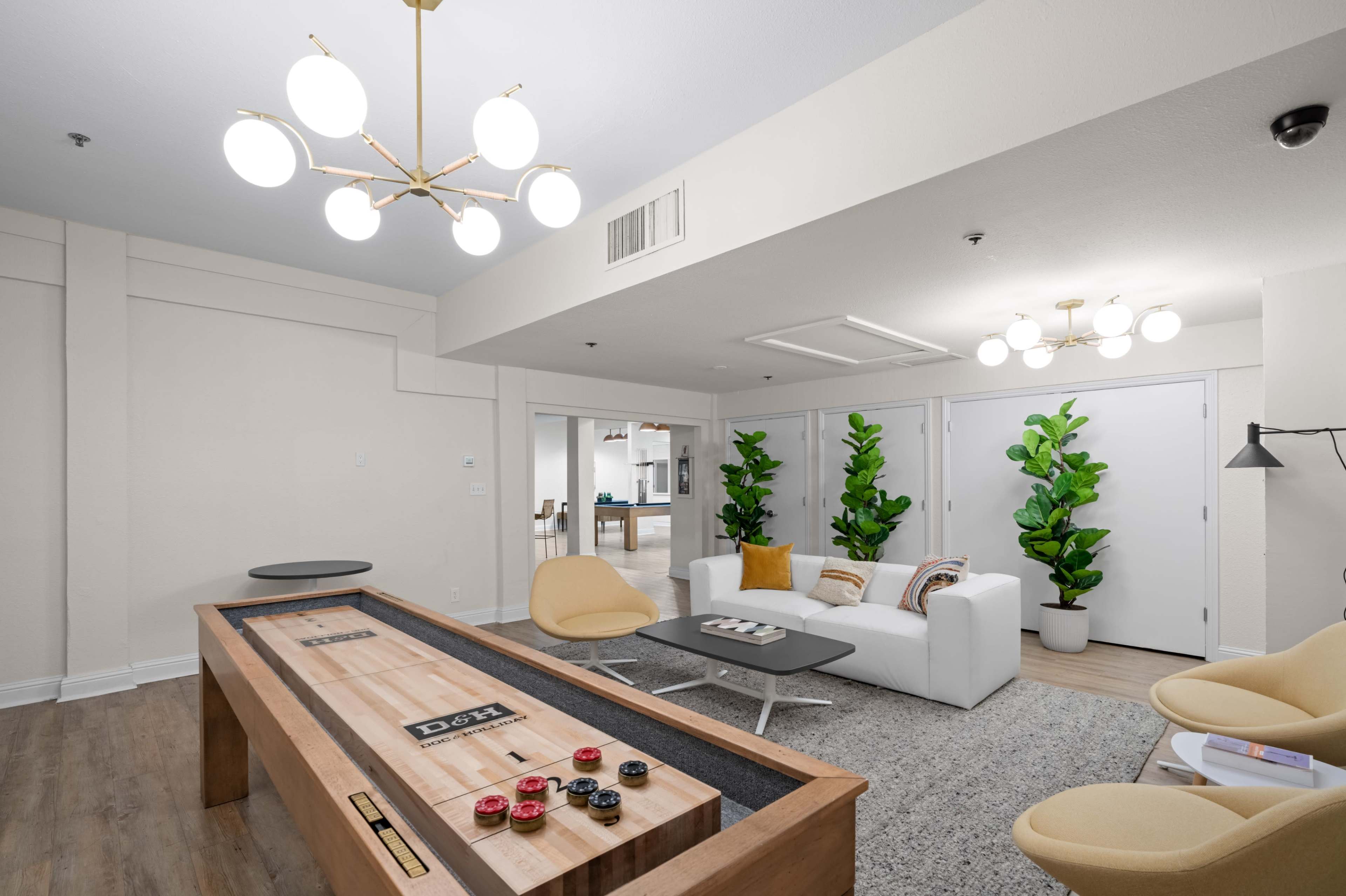 The image shows a modern lounge area featuring a shuffleboard table, a white sofa, two chairs, potted plants, and contemporary light fixtures.