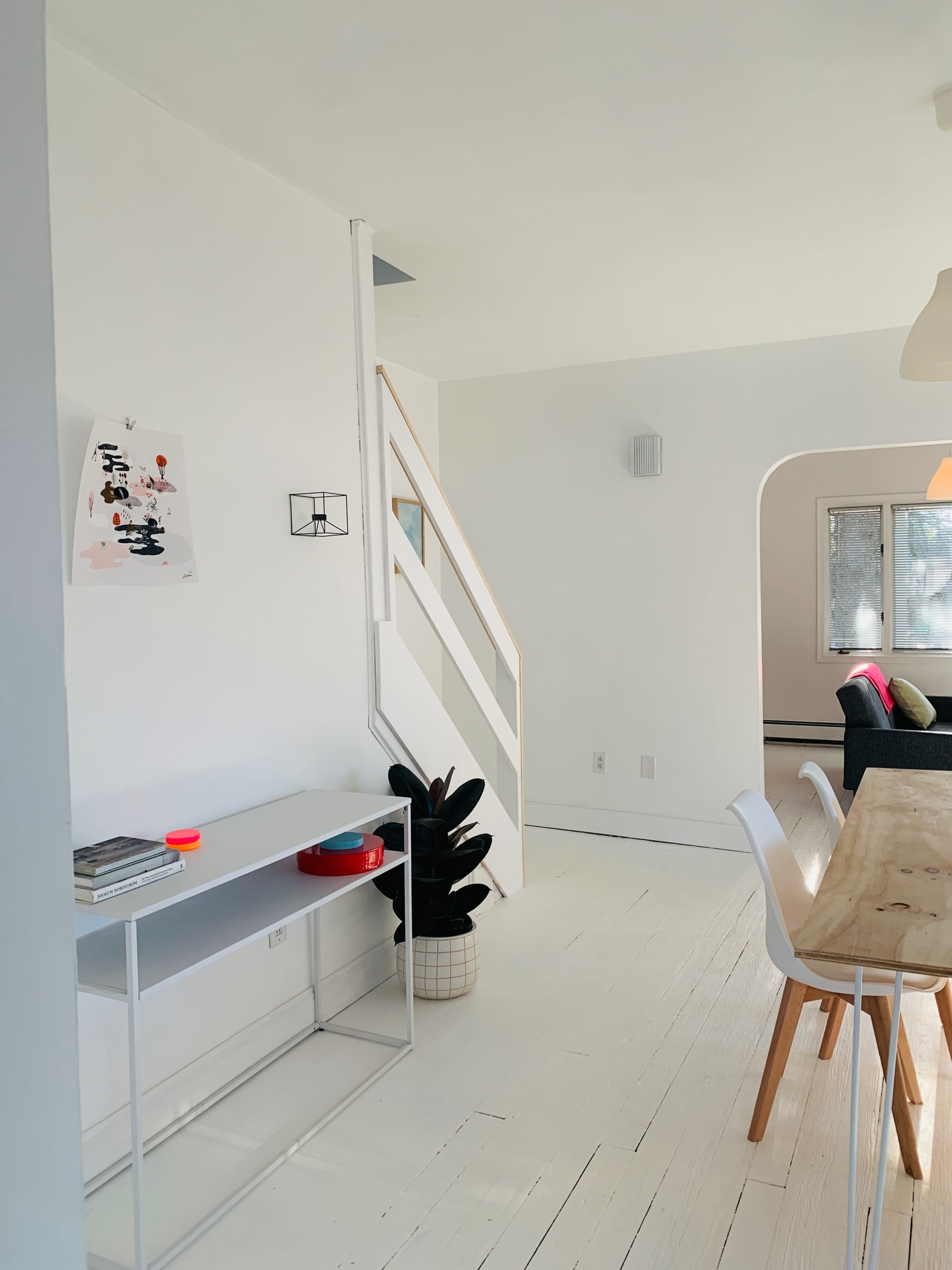 The image depicts a bright, minimalist interior of a room featuring a white wall and wooden staircase, with a simple table and chairs set up near a plant.