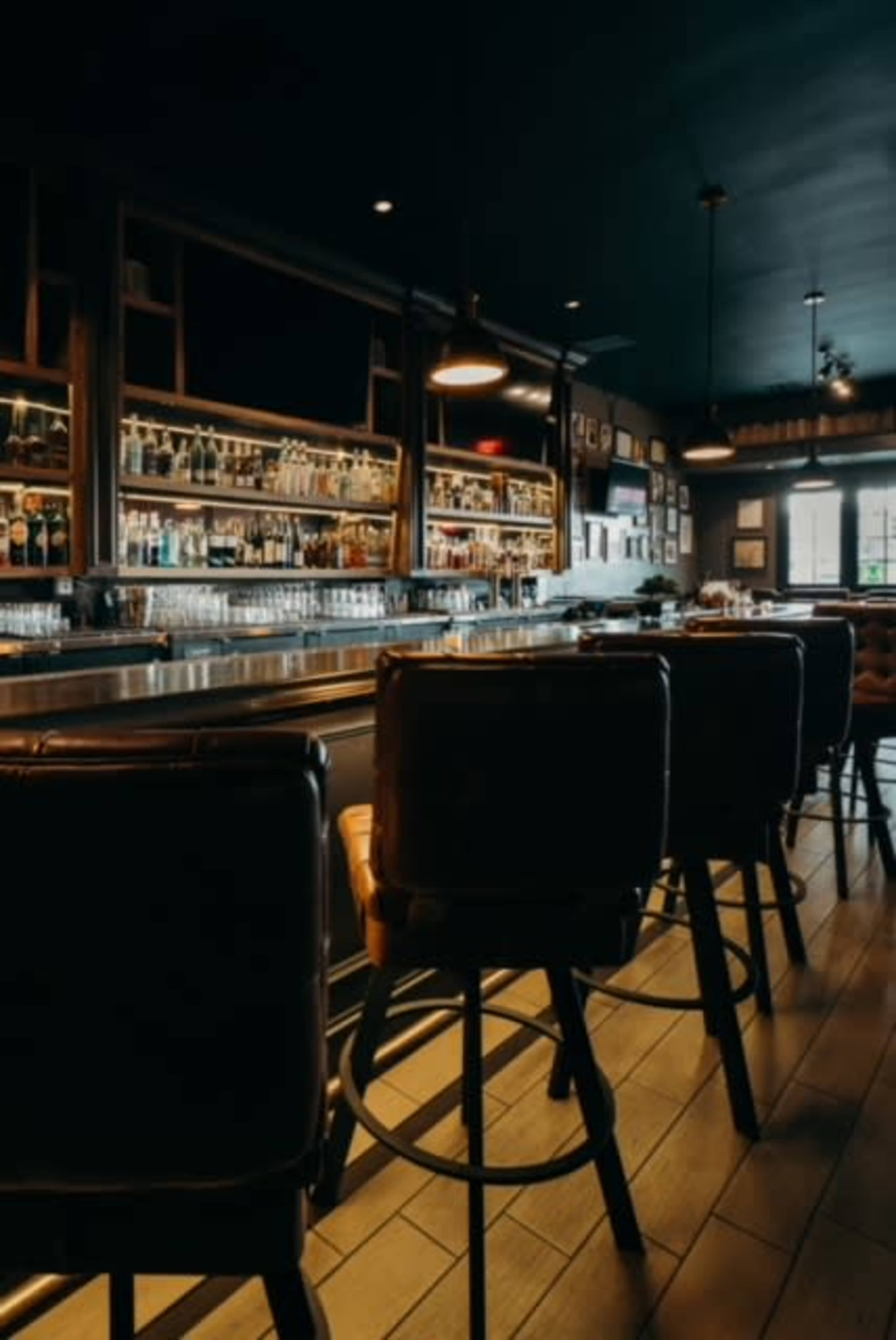 A dimly lit bar features a long counter with high stools and shelves lined with various bottles.