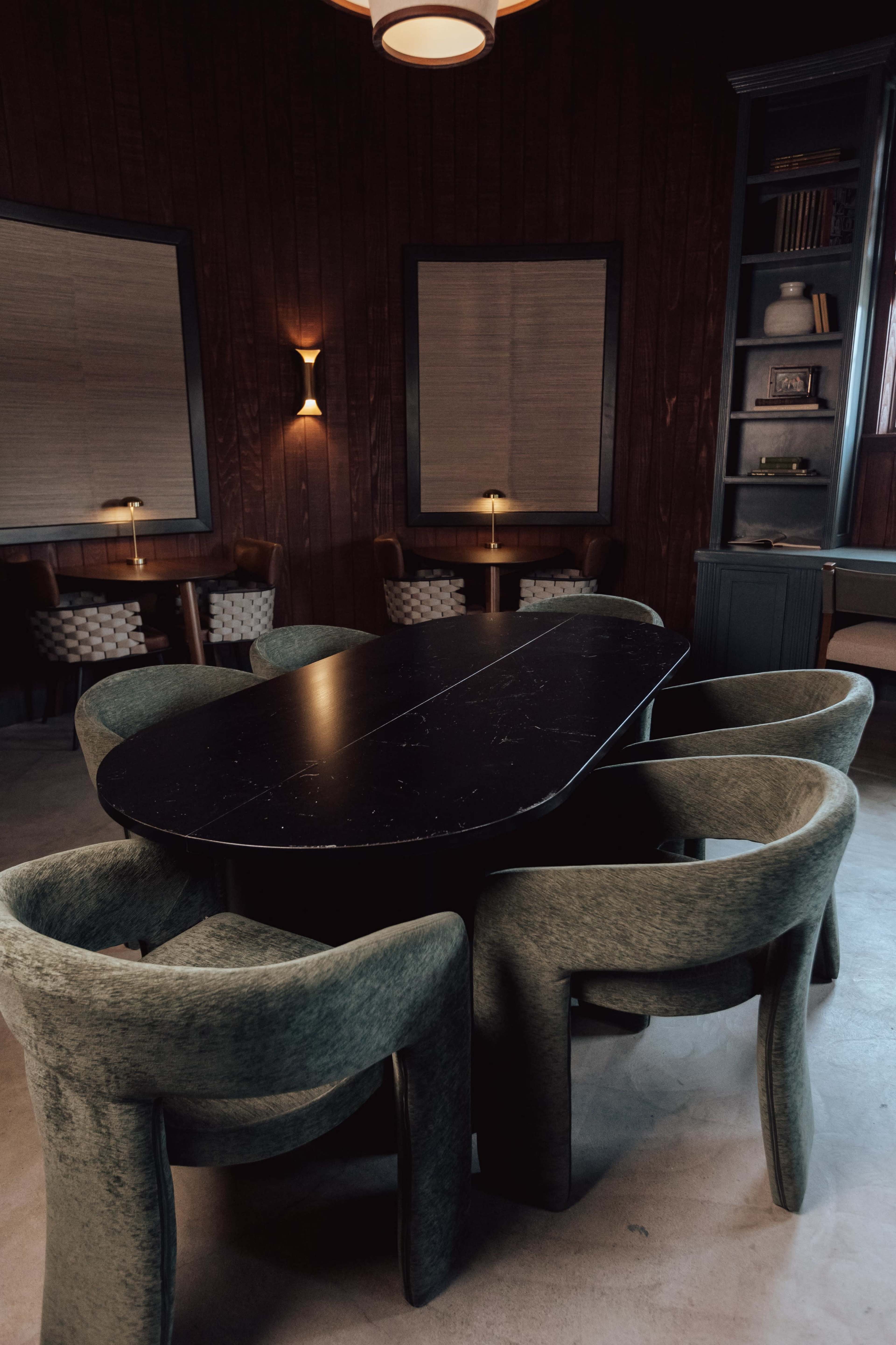 A round black table surrounded by green upholstered chairs sits in a dimly lit room with wooden walls and two wall-mounted lamps.