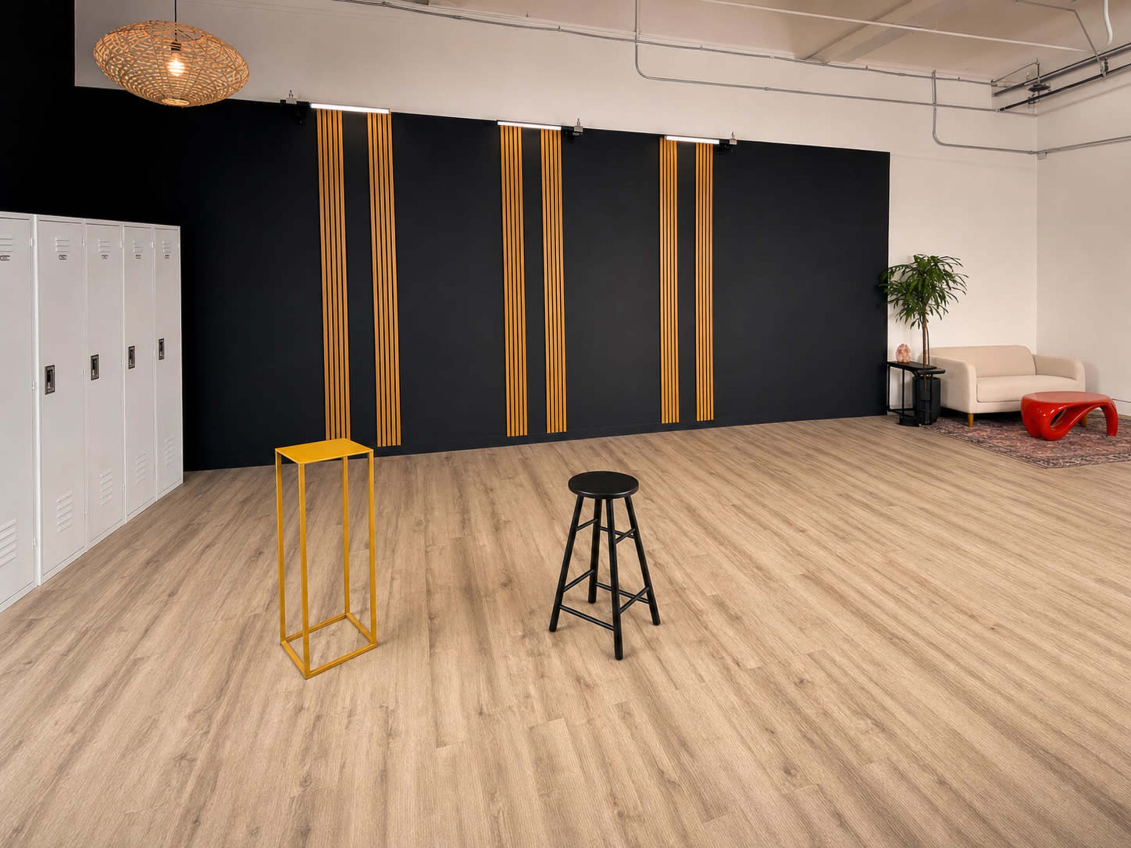 The image shows a spacious room with light wooden flooring, black walls featuring vertical gold stripes, a black stool, a yellow metal stand, lockers, and a white sofa.