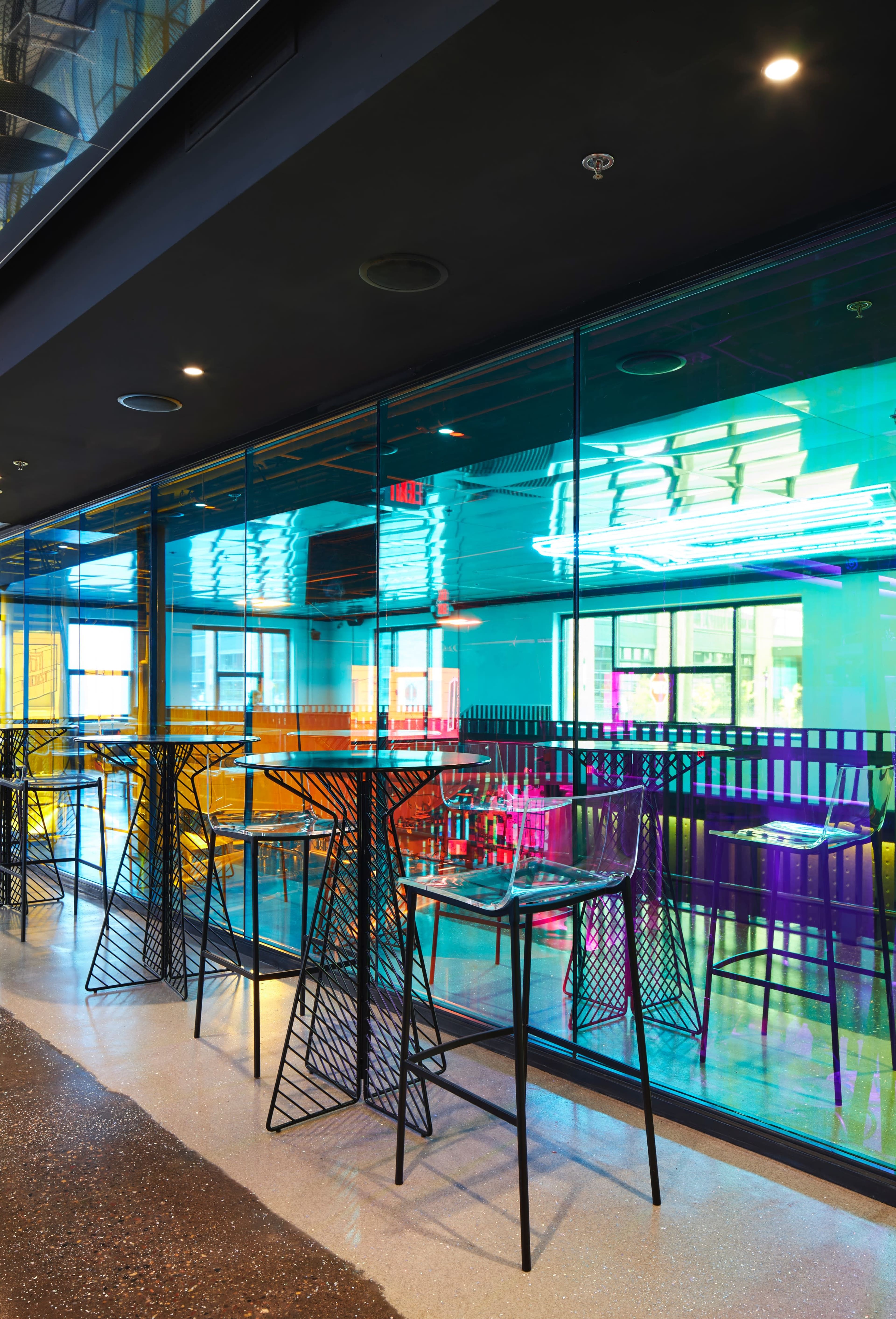 A modern cafe with colorful illuminated walls and transparent glass panels, featuring high black tables and chairs arranged in a spacious layout.