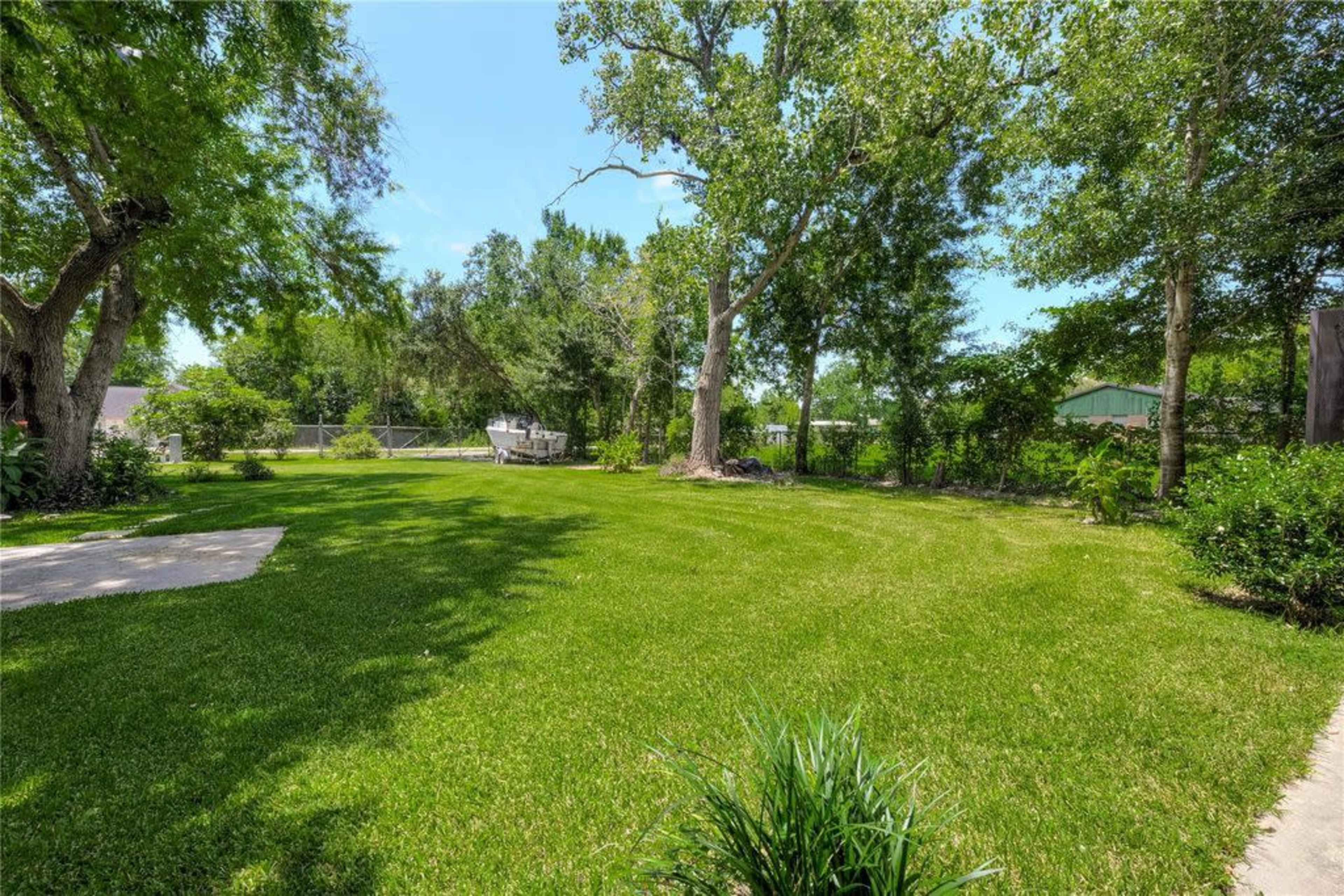 A spacious green lawn is bordered by trees and shrubs, with a clear sky and visible structures in the background.