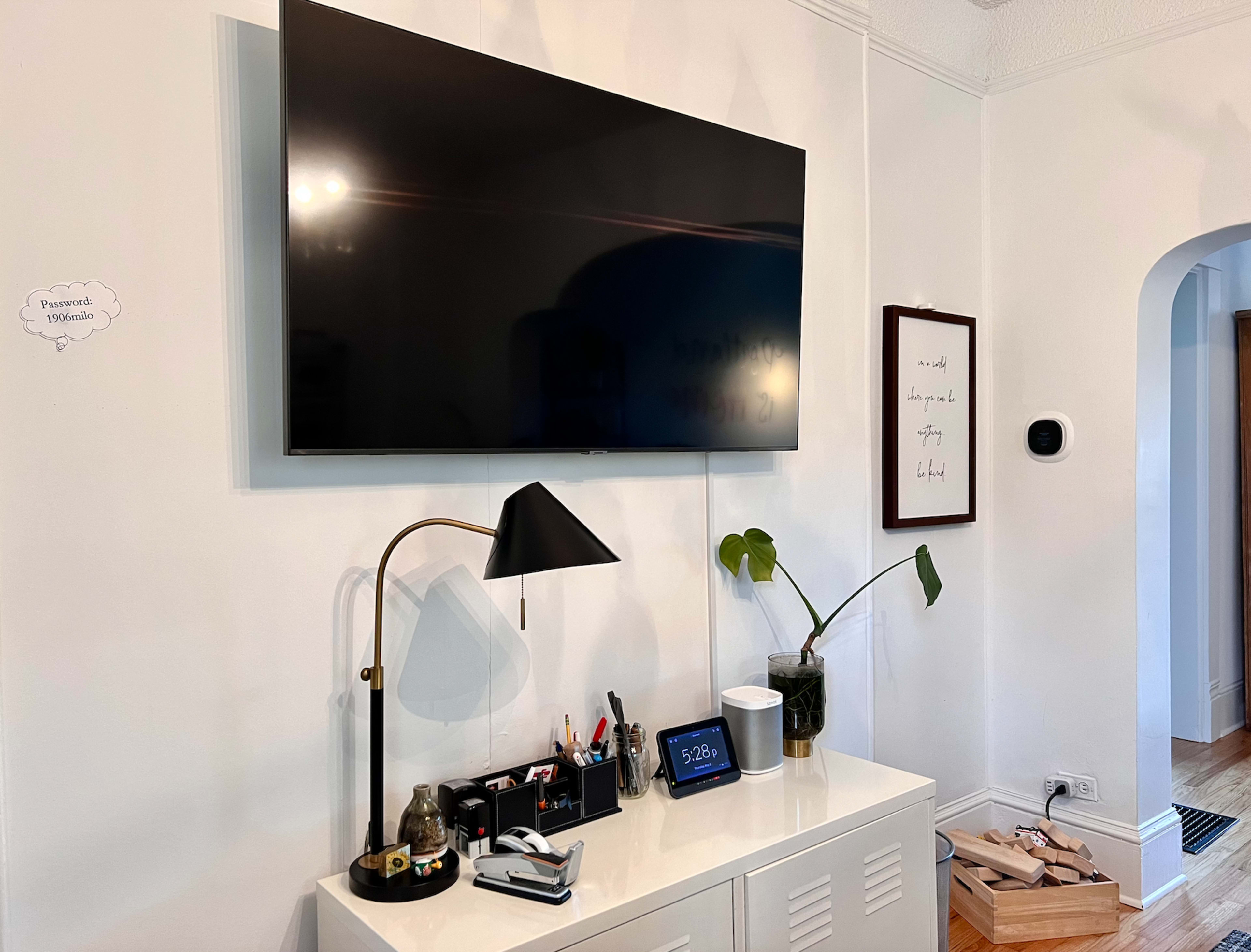 The image shows a mounted flat-screen TV above a white cabinet, with a desk lamp, various office supplies, a tablet, and a potted plant on the cabinet's surface.