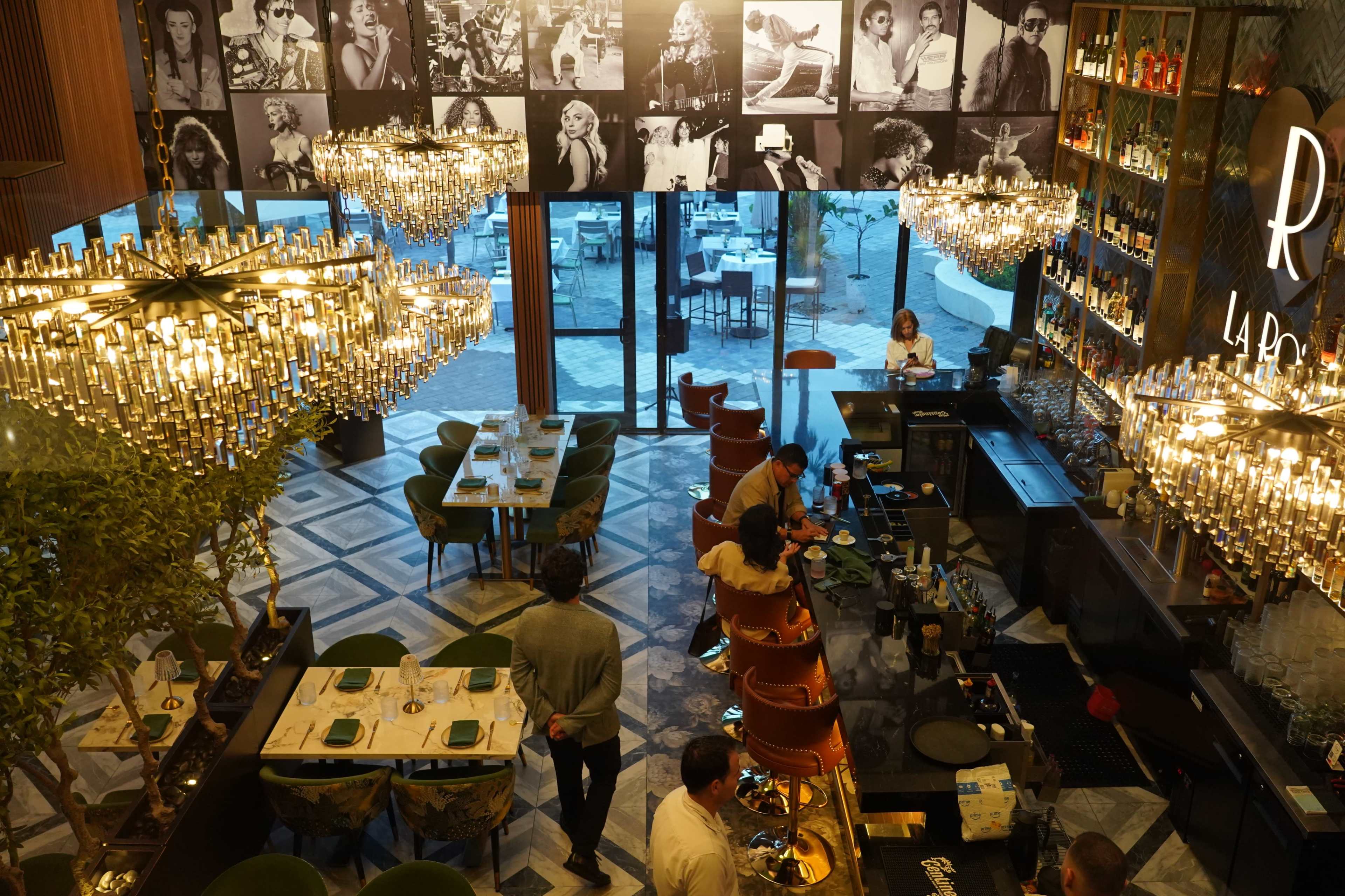 The image shows a stylish restaurant interior featuring a bar area, elegant chandeliers, and a collection of black-and-white portraits on the wall.