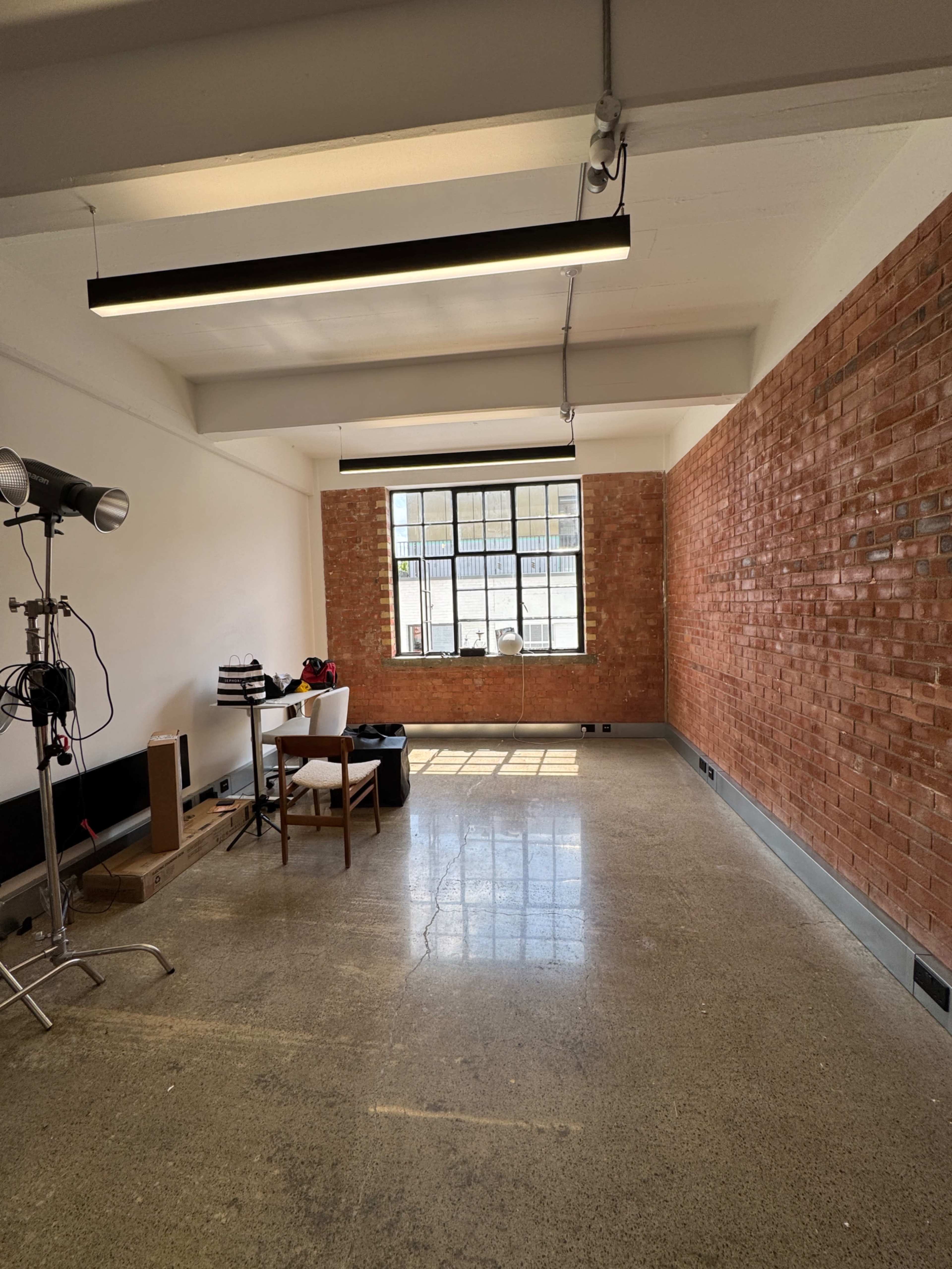 The image shows a spacious, empty room with a brick wall, a large window, a white table, a chair, and lighting equipment.