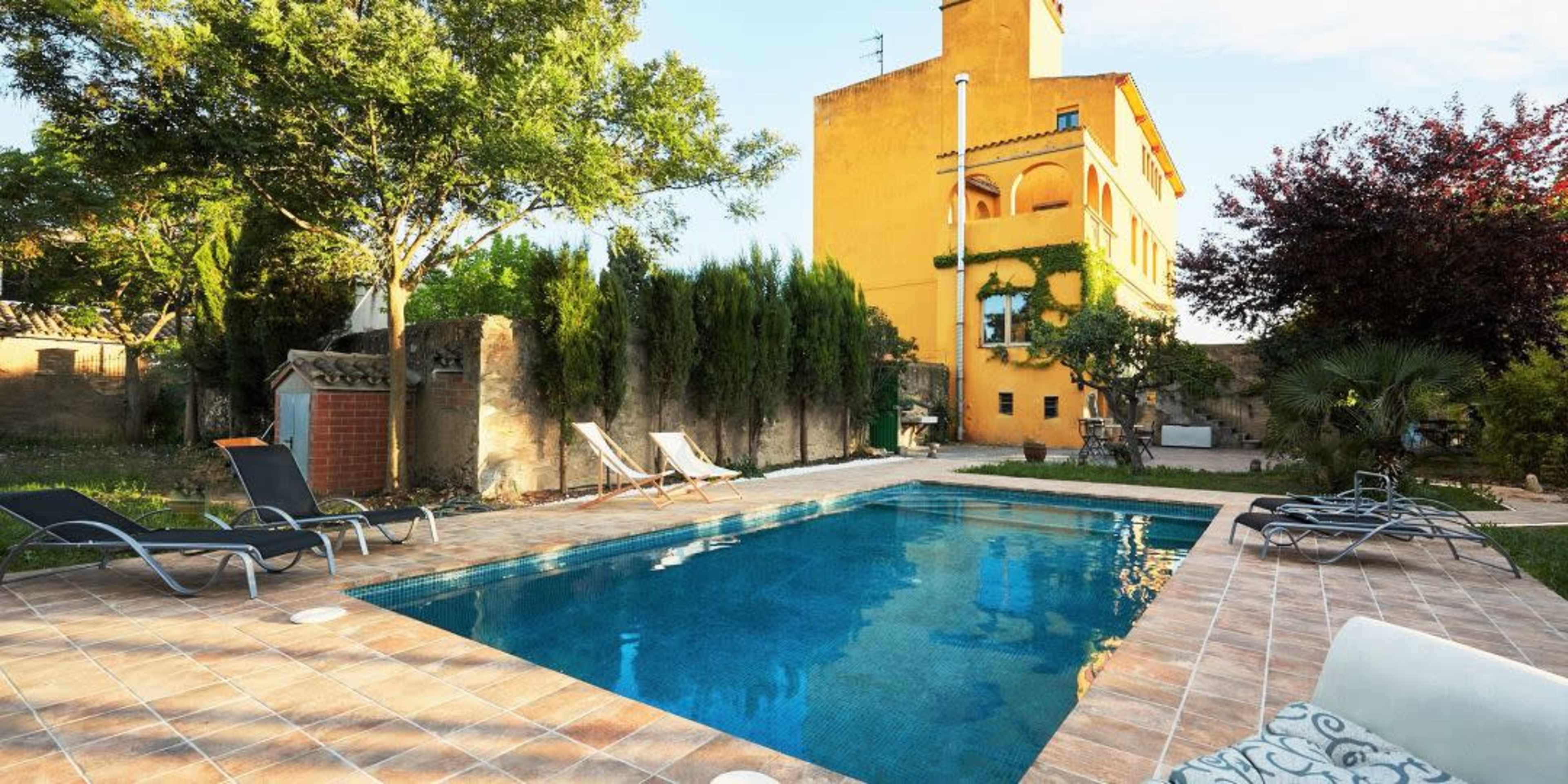 A rectangular swimming pool is surrounded by lounge chairs and greenery, with a yellow building in the background under a clear sky.