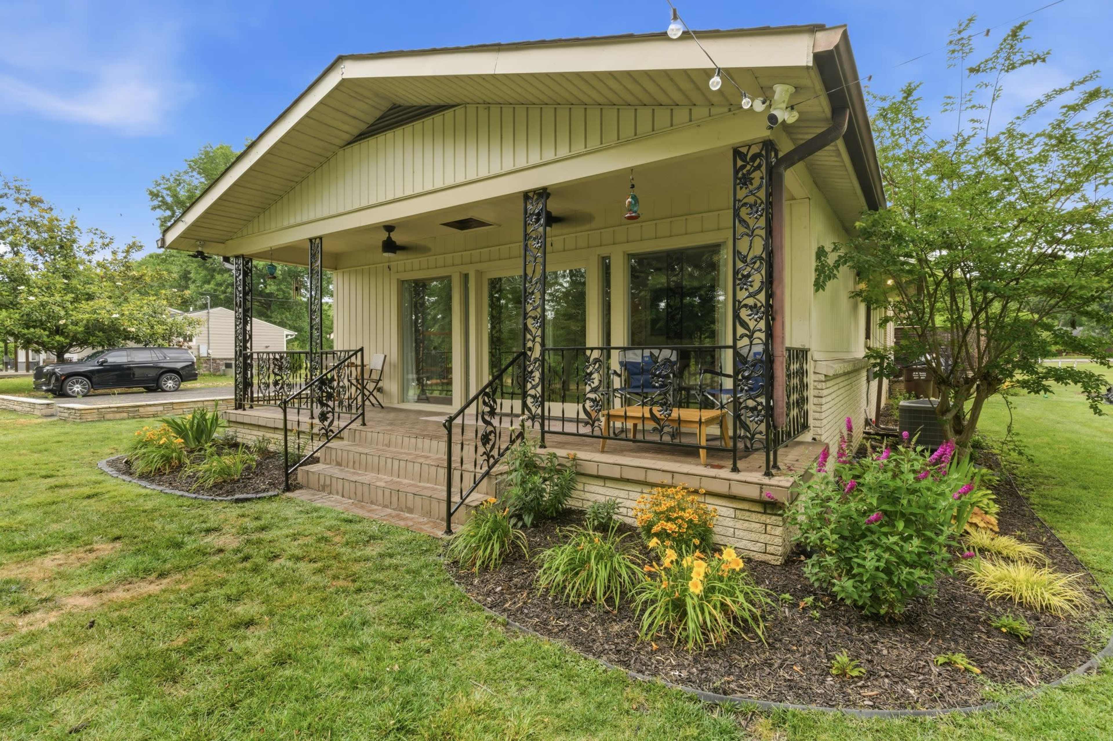 The image shows a house with a front porch surrounded by a landscaped yard featuring flowers and shrubs.