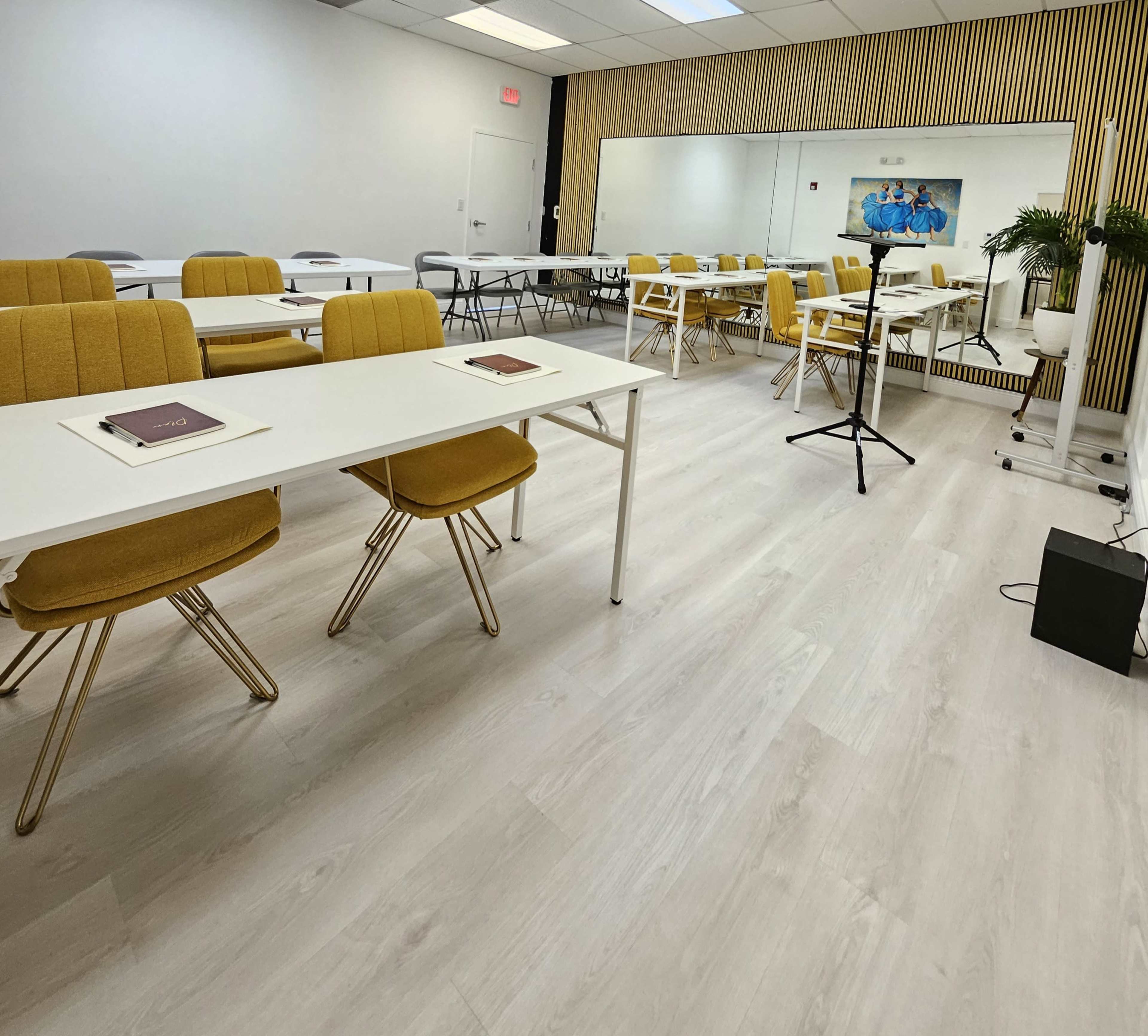 A bright, modern classroom setup with yellow chairs arranged around white tables and a large mirror on one wall.