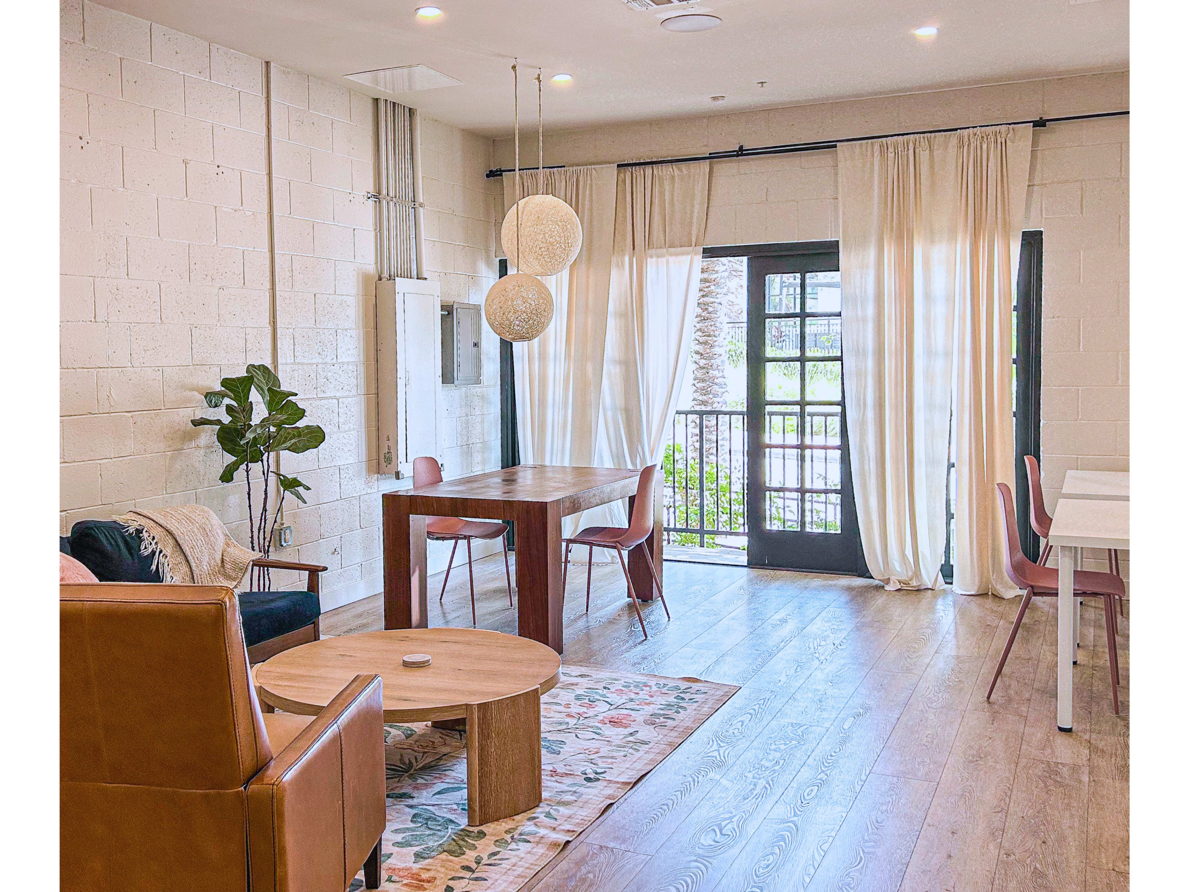 The image shows a modern living space with a wooden table and chairs, a sofa, and large windows allowing natural light to enter.