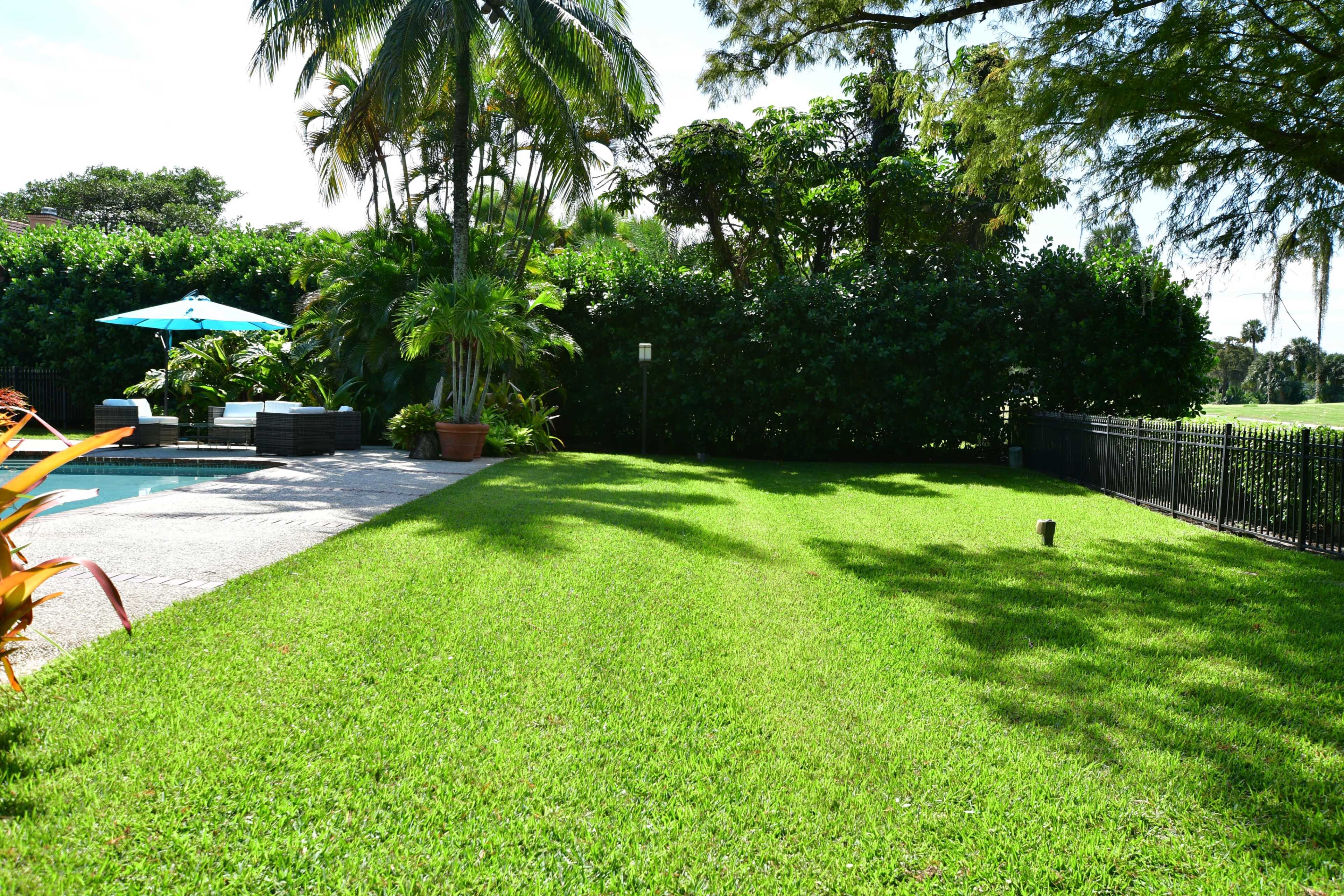 A well-manicured lawn stretches beside a swimming pool, flanked by tropical plants and a patio area with a blue umbrella.