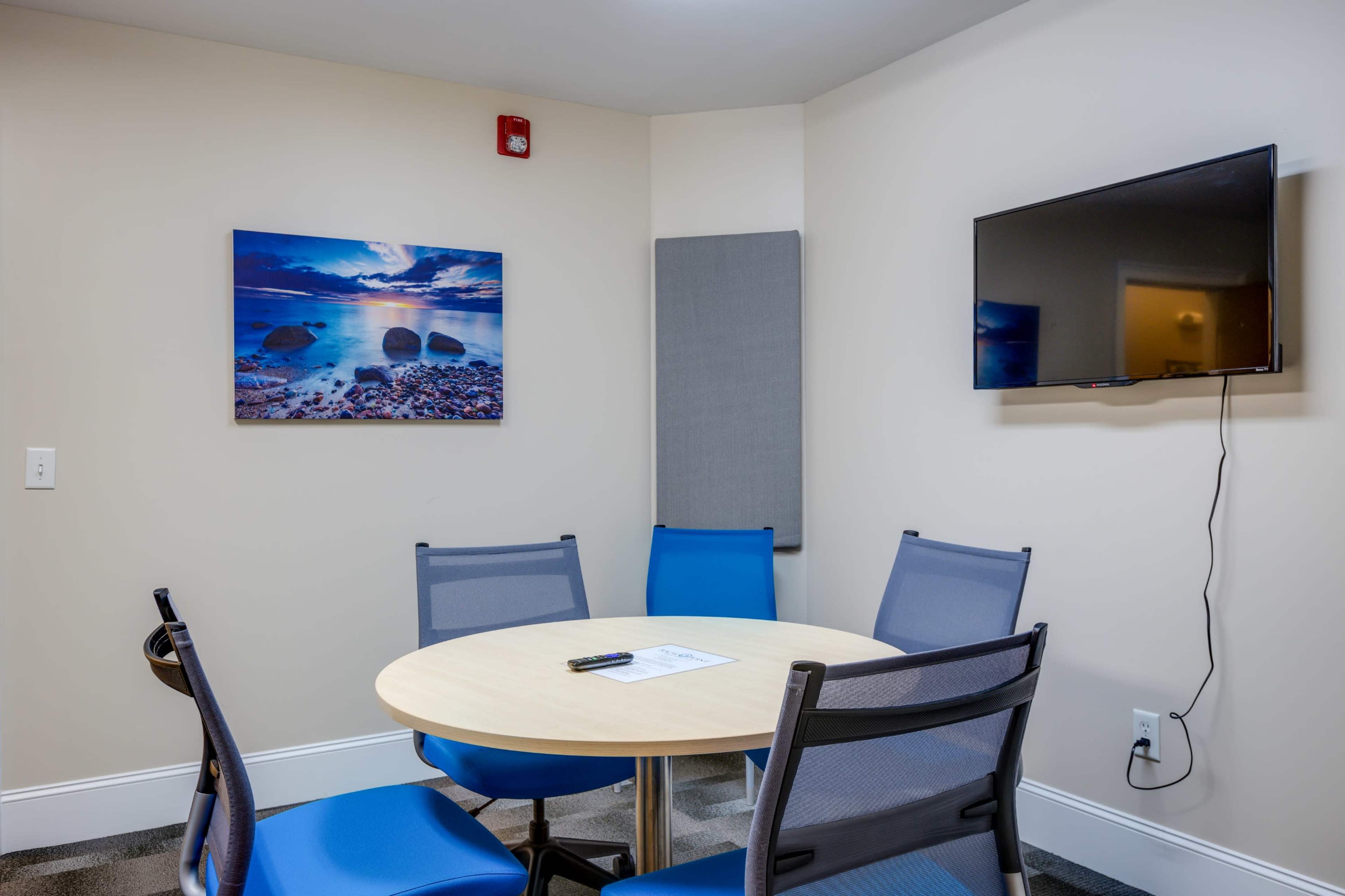The image shows a small meeting room featuring a round wooden table surrounded by blue chairs, a television mounted on the wall, and a landscape photograph.