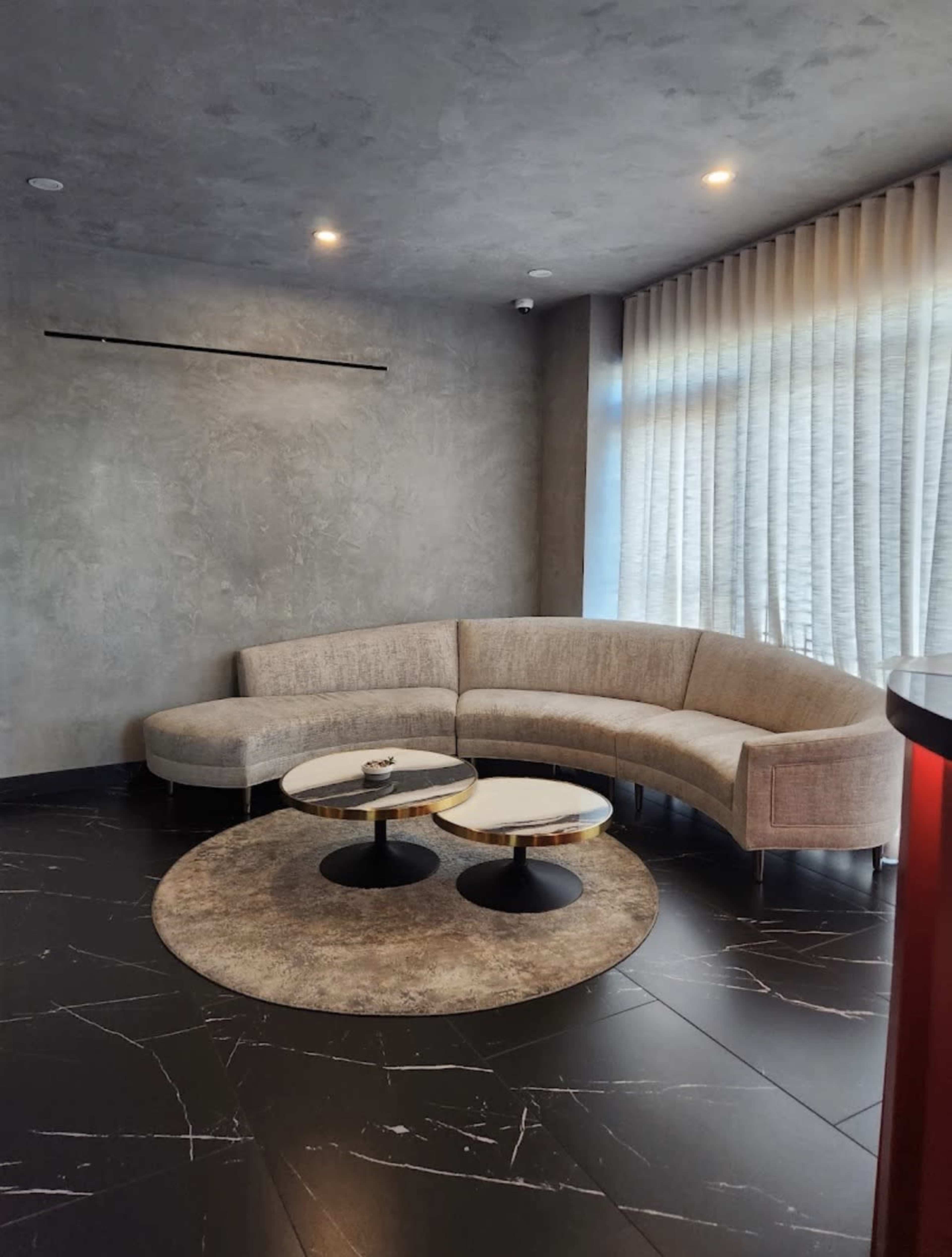 The image shows a modern lounge area featuring a curved sofa, two round tables, and a textured wall with large windows covered by sheer curtains.