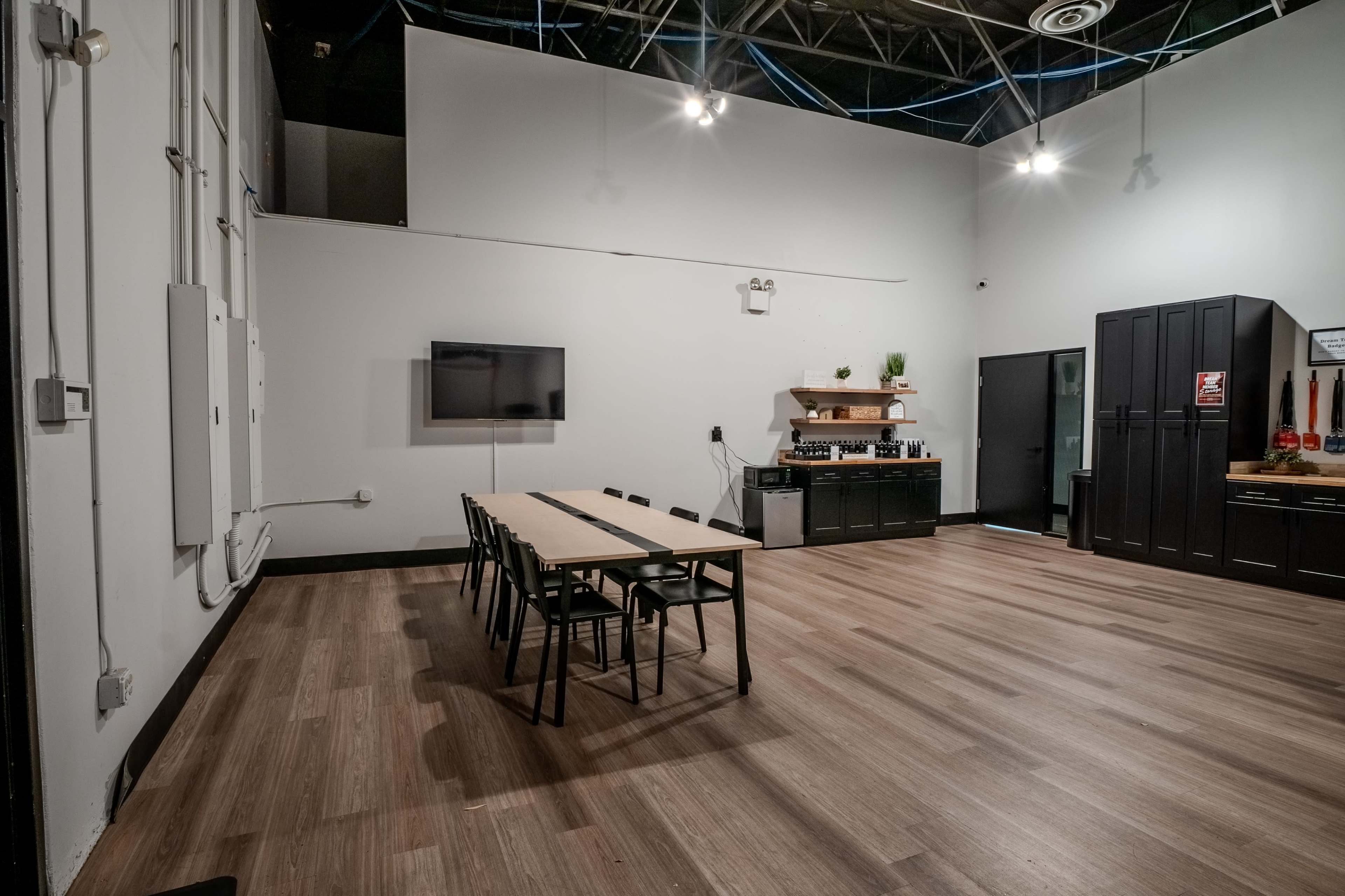 The image shows a large, light-colored room with a long table and chairs, a wall-mounted television, and a kitchenette along one side.