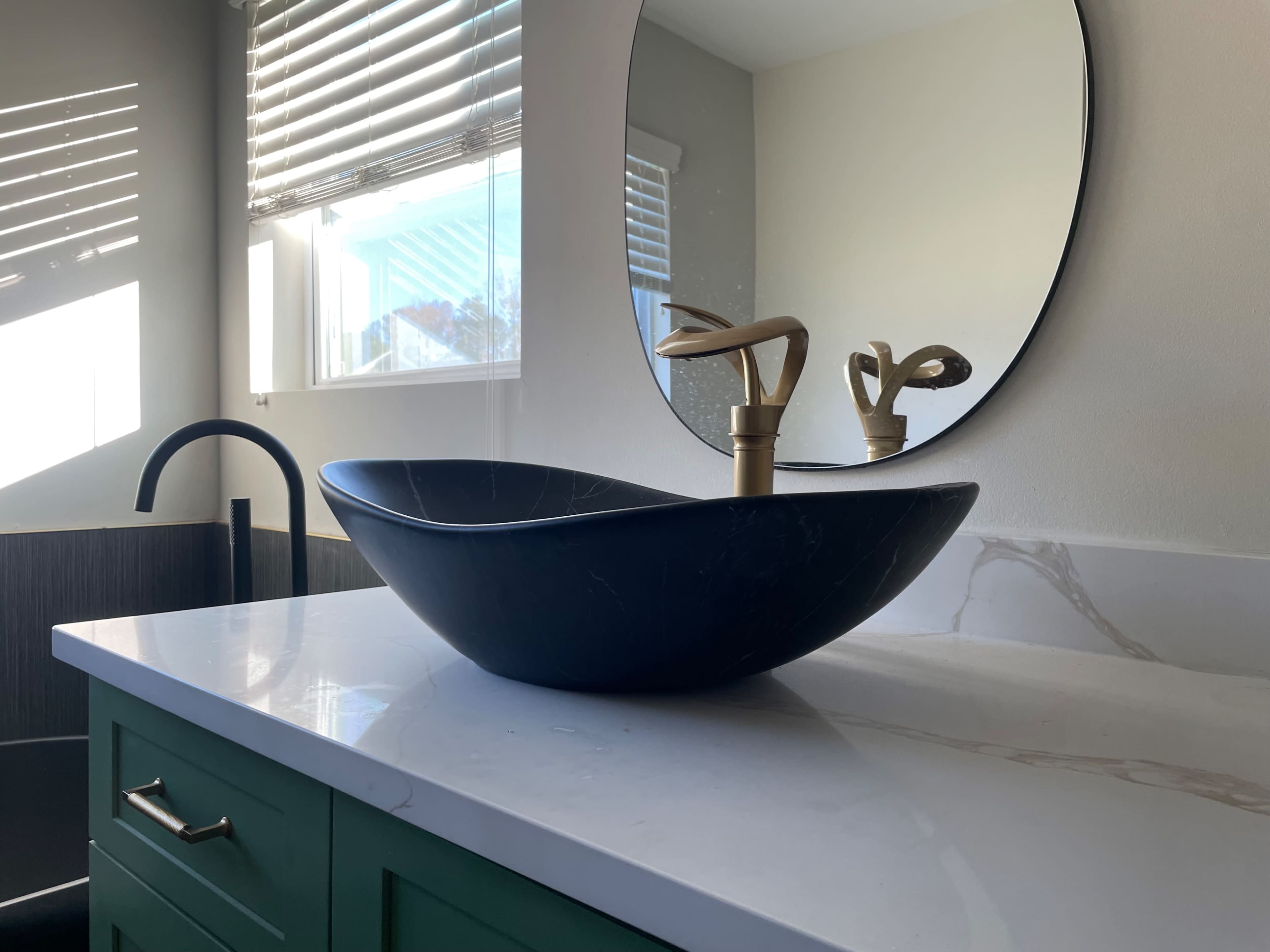 The image shows a modern bathroom sink made of black marble, positioned on a white countertop under a round mirror with natural light streaming in from a nearby window.
